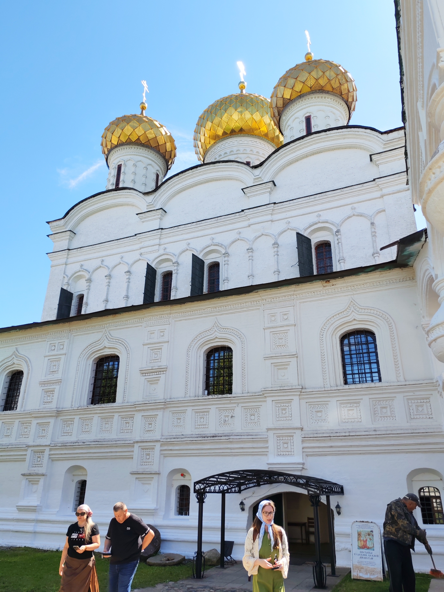 This image shows the Assumption Cathedral in Kostroma, Russia, under a clear blue sky. The cathedral is a large, white building with golden domes topped with crosses. The facade is ornate, featuring arched windows, decorative moldings, and a series of smaller arched niches. Several people are visible in the foreground. On the left, a woman wearing a black t-shirt, brown skirt, and sunglasses holds a phone. Beside her, a man in a black t-shirt and blue jeans looks down at something in his hands. Further to the right, a woman in a green dress and headscarf stands near an entrance. To the far right, a man in a camouflage jacket and cap sweeps the ground with a broom. The ground is a mix of grass and paving stones, and a black metal awning shelters the entrance to the building. A sign with text and an image is also visible near the man sweeping.