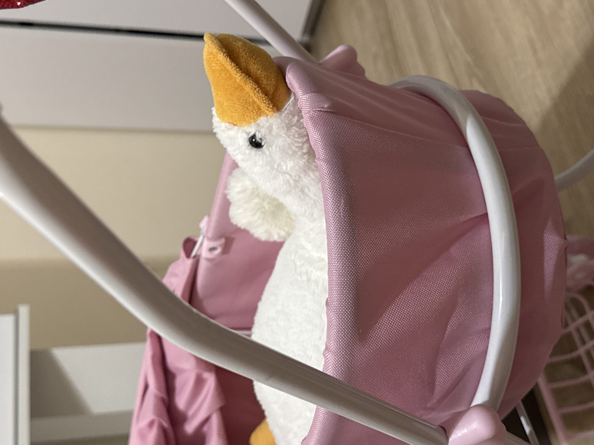 The image shows a close-up of a stuffed duck toy partially covered by a pink fabric canopy, which appears to be part of a toy stroller or bassinet. The duck has white, fluffy fur and a yellow beak. Its small black eye is visible. The fabric is a soft pink and has a textured weave. White, curved bars of the toy's frame are prominent in the foreground, framing the duck and canopy. In the background, there are hints of a room with neutral-colored walls and furniture, suggesting an indoor setting. The lighting appears to be artificial, casting soft shadows. The overall impression is of a child's toy or nursery item. There is no visible text. The location context is Mikhaylovsk, Russia, though no specific visual cues in the image directly confirm this.