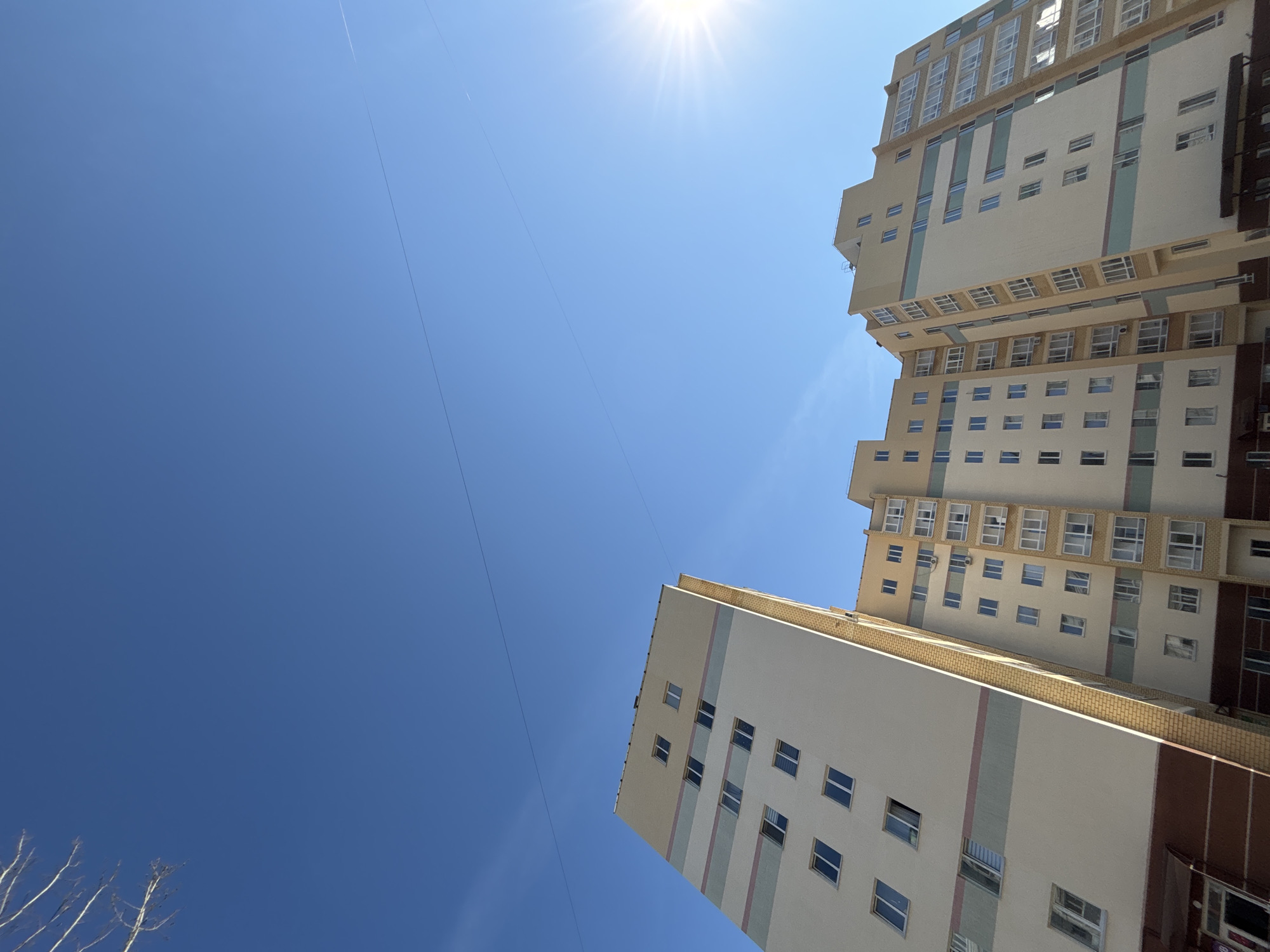 The image shows two tall buildings under a clear blue sky with the sun shining brightly. The buildings appear to be residential, with multiple windows on each floor. The architectural style is modern, with a mix of white, beige, and brown colors on the facade. The buildings are located in the city of Chaikovsky, Russia. A thin, straight line crosses the sky, possibly a contrail from an airplane. Some bare tree branches are visible in the lower left corner.