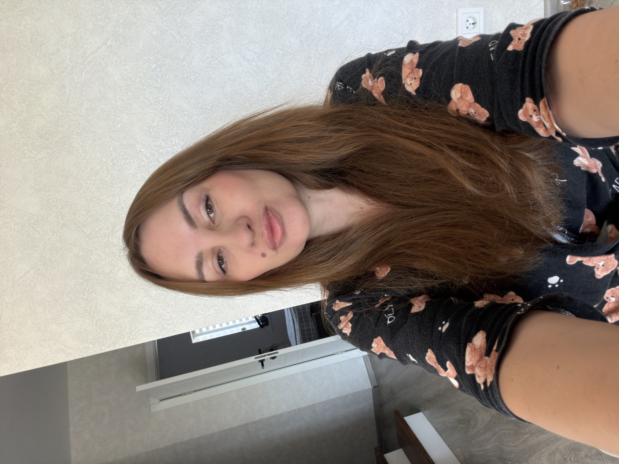 A young woman with long brown hair and a mole on her cheek poses for a selfie in a room. She is wearing a black long-sleeved shirt with a pattern of teddy bears. The background shows a door, a staircase, and a textured wall with a power outlet. The lighting is bright, indicating it's daytime.