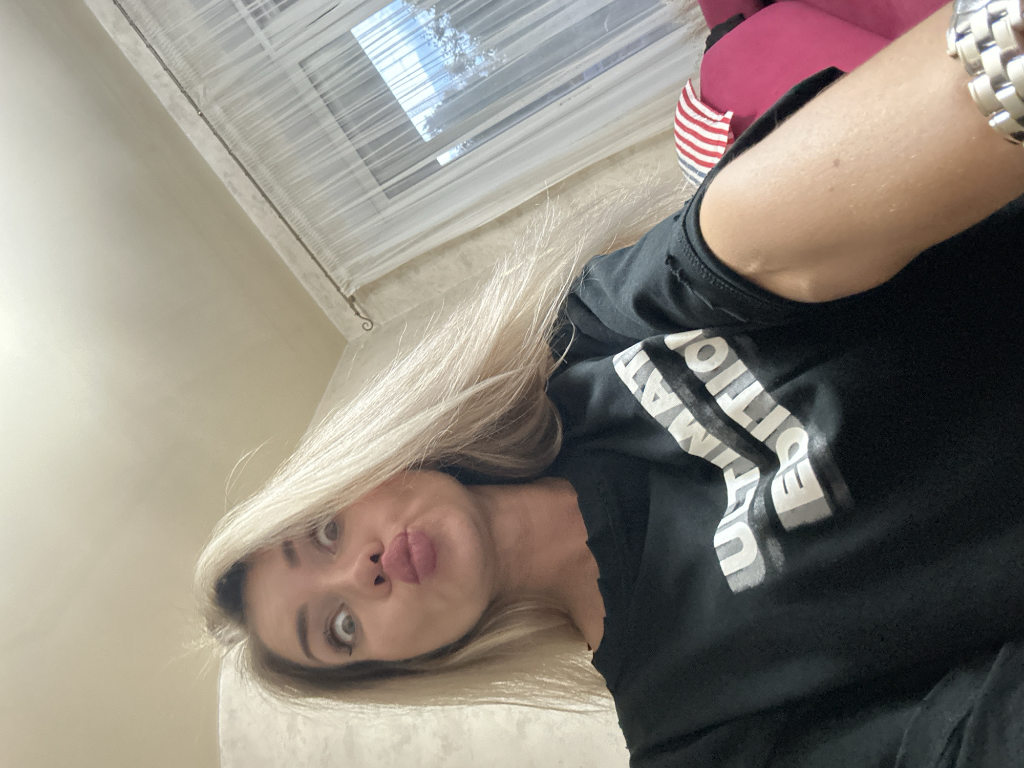 A young woman with blonde hair pulled into a messy bun and wearing a black t-shirt with the words "ULTIMATE EDITION" is captured in a selfie, puckering her lips and widening her eyes in a playful expression. Her head is tilted, and she is looking directly at the camera. The background shows a neutral-colored ceiling, sheer white curtains covering a window, and a hint of a pink couch with a red and white striped pillow. The lighting appears to be natural, coming from the window. The setting suggests an indoor residential environment. The location of Armavir, Russia is not determinable from the image.