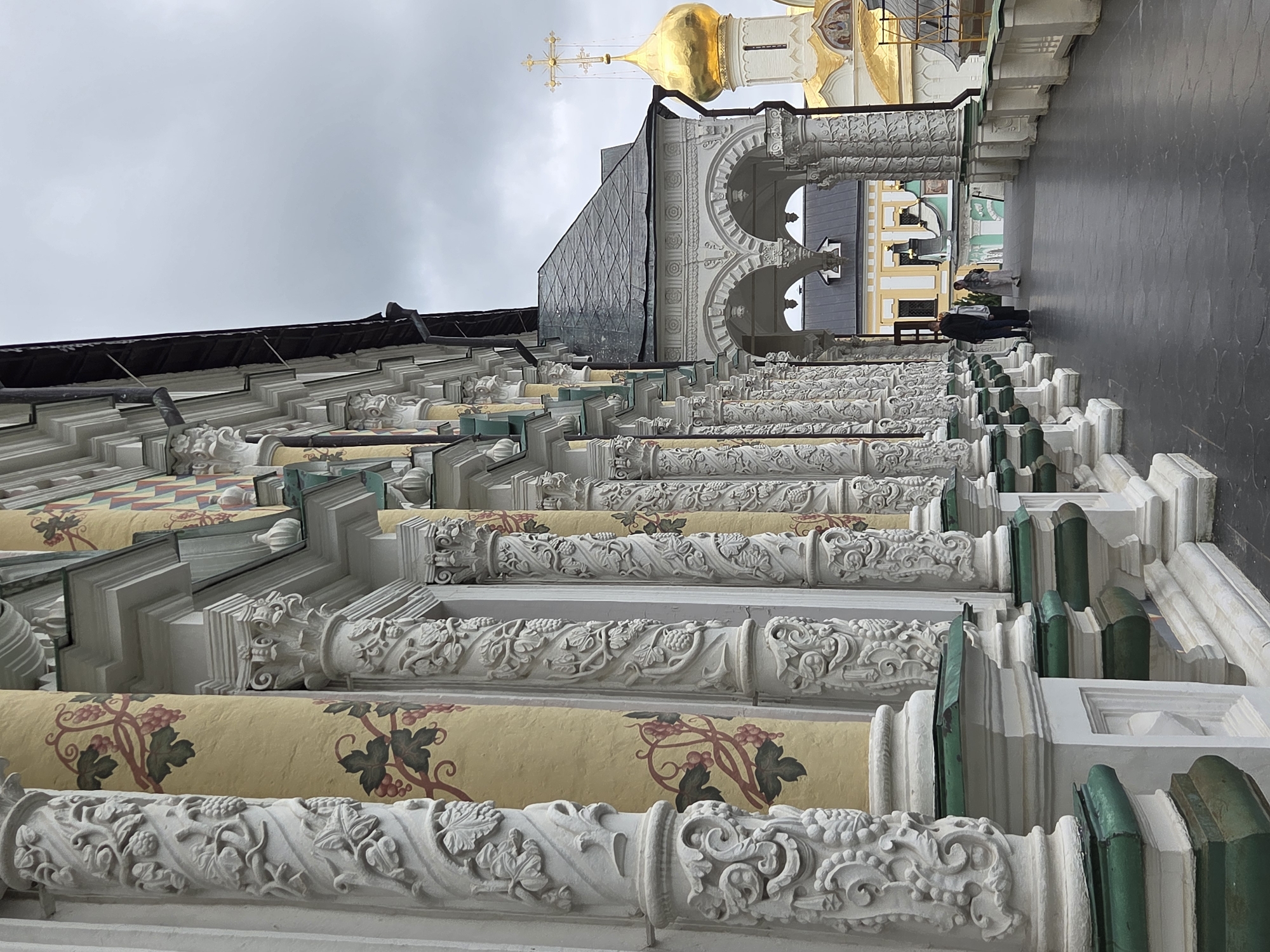 The image captures a detailed view of the Trinity Lavra of St. Sergius in Sergiev Posad, Russia. The foreground features a row of intricately carved white columns adorned with floral and grape motifs. These columns are set against a yellow facade painted with a vine and grape pattern. The architectural style is ornate, with detailed cornices and decorative elements. A dark gray, paved walkway runs alongside the columns, suggesting an outdoor covered passage. In the mid-ground, two figures, a man and a woman, are walking away from the camera. Beyond them, an arched walkway leads to buildings with a golden dome topped with a cross, characteristic of Orthodox churches. The sky is overcast and gray. The overall scene depicts a historical and religious complex with rich architectural detail and a somber atmosphere due to the cloudy weather.
