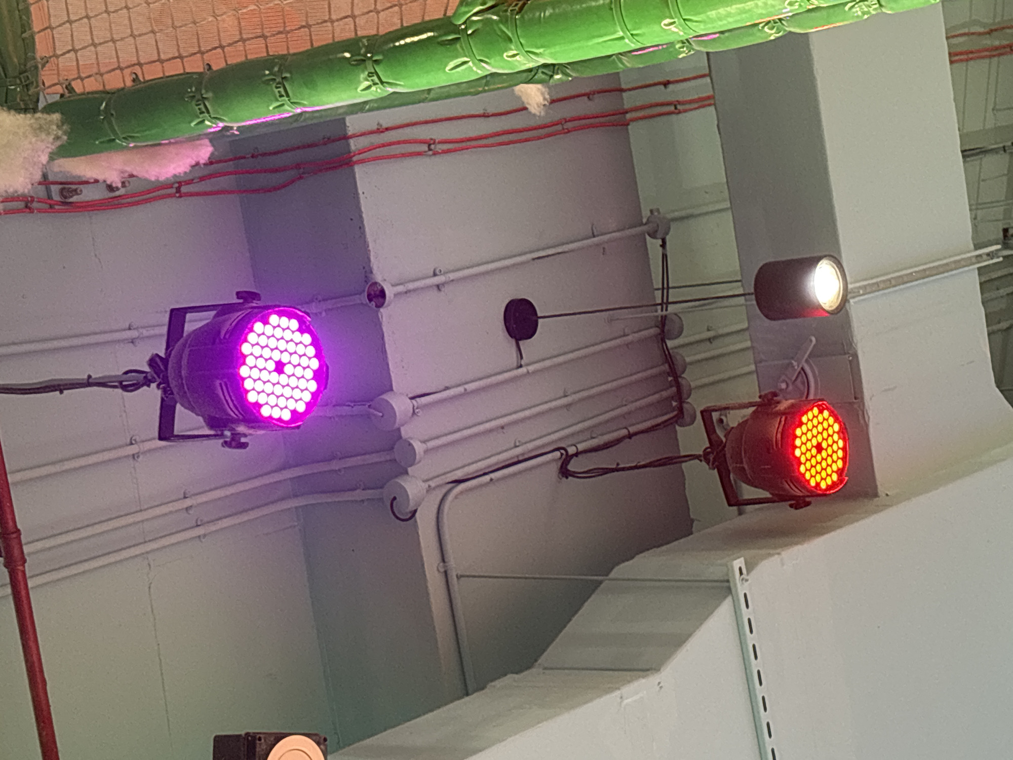 This is a close-up, eye-level, medium shot of the interior ceiling area of a building, likely an entertainment venue or a space with stage lighting, as evidenced by the three spotlights. The lighting setup includes one large, purple LED spotlight with a grid of lights, hanging from a black metal bracket. Below and to the left, a red LED spotlight is visible, also with a grid of lights, and appears to be in a darker, possibly metallic housing. To the right of the red spotlight, and further to the right and slightly down from the purple one, is a cylindrical pendant light with a white bulb illuminating downwards. The ceiling is a light gray, with various conduits and electrical wiring running across it. A thick, green, cylindrical structure, possibly a padded pillar or a decorative element, occupies the right side of the frame, with red wires running alongside it. There is no indication of the specific location being Dubai, United Arab Emirates.
