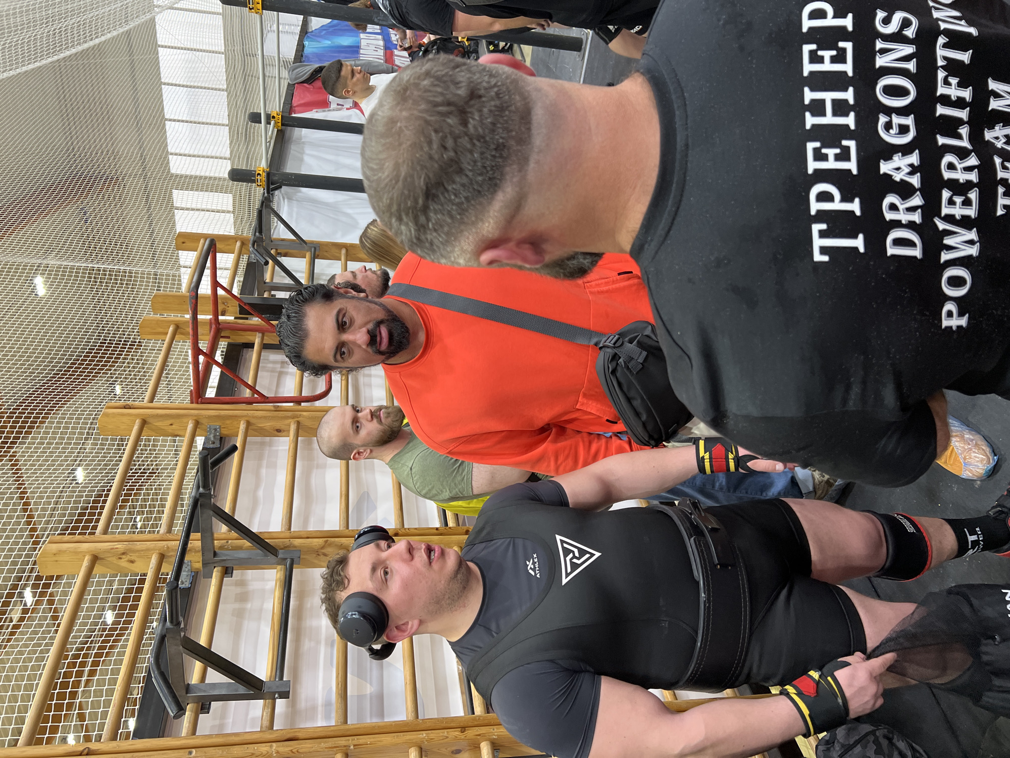 The image shows an indoor scene, likely a gymnasium or training facility. A weightlifter, wearing a black suit and wrist wraps, is lying on his back. He has headphones on his head. Several people surround him. One individual, wearing an orange sweatshirt and a black shoulder bag, is looking down at him. Another person, wearing a black t-shirt with the words "ТРЕНЕР DRAGONS POWERLIFTING TEAM", is standing nearby. In the background, there's a wooden climbing frame and a safety net. Based on the team name written in Cyrillic, this event is likely occurring in Russia. Without additional information from the image, I can not guarantee the exact location of the city, Saint Petersburg.