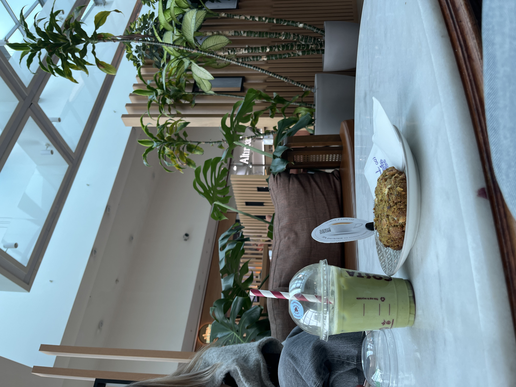 Here is a forensic description of the image:
**Location:** An interior setting, likely a café or restaurant, located in Sofia, Bulgaria. The brand Altır Ja is visible in the background.
**People:** A partially visible person is seated at the table, wearing grey clothing (sweater/cardigan and denim jeans). Only the hair of the person, clothing, and legs are visible.
**Objects:**
* A marble table is featured prominently.
* On the table, a green matcha drink is in a transparent plastic cup with a red and white striped straw.
* A plate containing a pastry item (muffin), accompanied by a napkin, is also on the table.
* A small coaster-like object with the words "espresso" and “отстъпк” is adjacent to the matcha and the plate with pastry.
* Plants are visible in the background and around the seating area.
* Potted plants.
**Environment:** The location appears to be indoors. The background features architectural elements such as ceiling windows and a wall with horizontal wooden slats.
**Actions/Interactions:** No specific action is taking place. The objects suggest someone is in the process of consuming or has recently consumed the drink and pastry.
**Additional Details:** The image is rotated, making it appear as if the table is vertical. The setting has elements of modern interior design with a focus on plants and a marble tabletop.