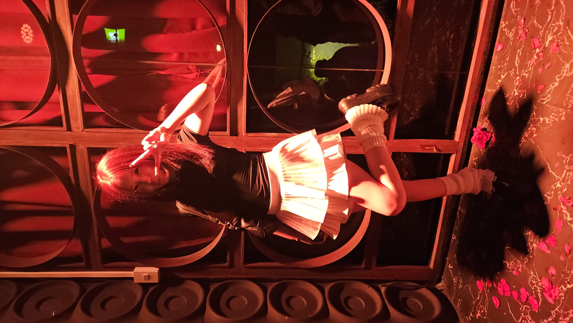 A person with red hair is positioned in front of a set of circular wall decorations. The person, wearing a black top, a white skirt, leg warmers, and platform shoes, is holding up two fingers. The scene is bathed in red light. The wall to the right has a floral pattern. The setting is in Nha Trang, Vietnam.
