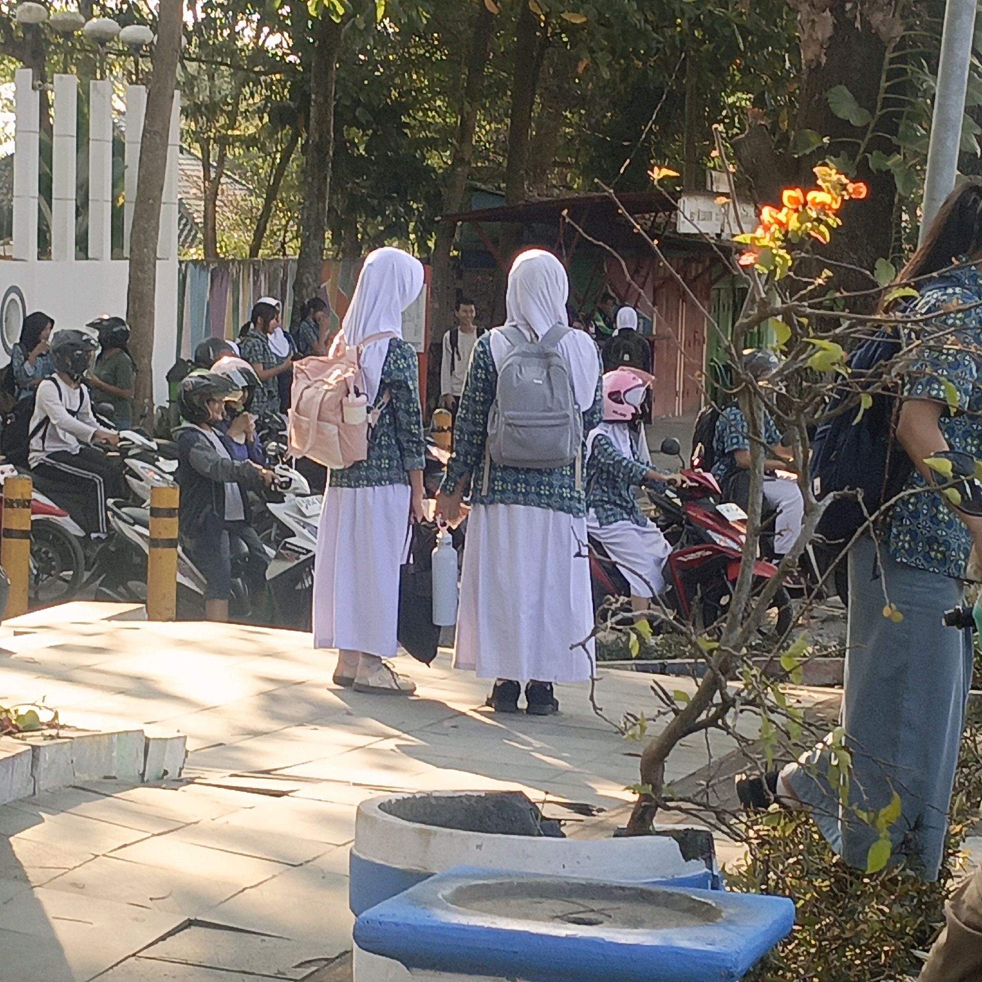 The image depicts a group of young people, likely students, gathered outdoors in what appears to be a school or community area in Salatiga, Indonesia. The sun is shining, casting long shadows on the paved ground. Many of the individuals are wearing hijabs and school uniforms, consisting of patterned shirts and long white skirts. Several are seen with backpacks.
In the foreground, two young women in uniform stand facing away from the camera, each with a backpack. One has a light pink backpack, and the other a gray one. To their left, a line of parked motorcycles is visible, with several people in helmets standing or sitting on them, suggesting a mode of transport. One individual in a white long-sleeved shirt and black pants is seated on a motorcycle. Another person, also in uniform, is standing next to a motorcycle, holding a grey water bottle.
Further back, other individuals can be seen in similar attire, some in conversation. A man in a light-colored shirt is visible standing near a wooden structure. Trees and foliage surround the area, with a flowering plant in the mid-right of the frame adding a touch of color with its orange and yellow blossoms. The overall impression is one of a bustling scene, possibly at the beginning or end of a school day. The presence of multiple motorcycles indicates a common mode of transportation for the students in this Indonesian setting.