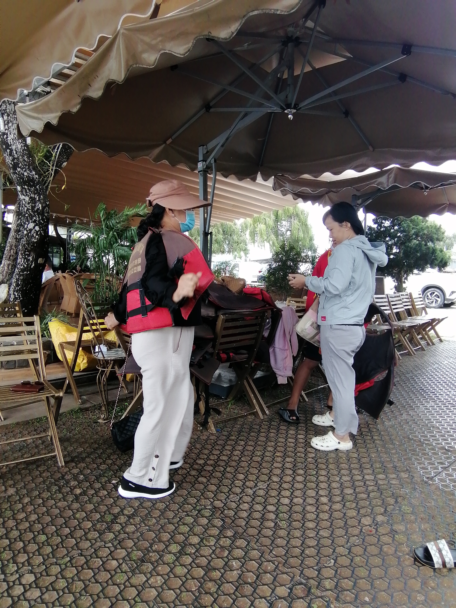 This image depicts an outdoor seating area under a large brown umbrella, likely at a cafe or restaurant in Da Lat, Vietnam. Two women are the primary subjects. The woman on the left wears a light pink hat, a light blue face mask, a dark jacket over a red and black vest with white lettering, and loose-fitting beige pants. She appears to be in mid-motion, possibly dancing or gesturing. The woman on the right, facing away from the camera, wears a light blue hooded jacket and light grey pants. She is holding a small object, possibly a phone, in her hand. Several empty wooden chairs and tables are visible, along with some potted plants and trees. The ground is paved with hexagonal stones. In the background, other tables and chairs are set up, and a white car is parked. The scene suggests a casual, possibly recreational, setting.