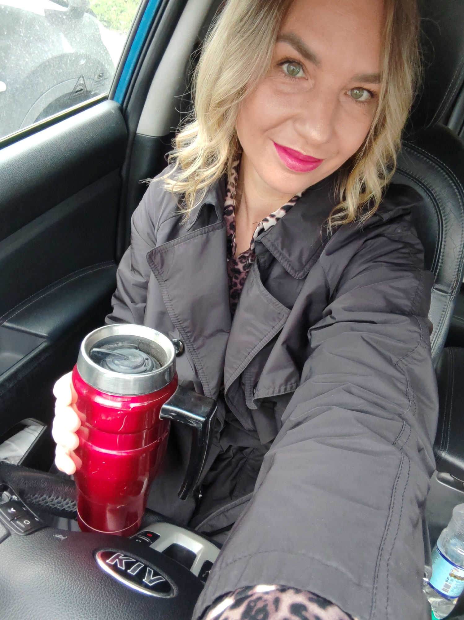 This image is a selfie taken by a woman inside a car, likely in Petropavlovsk-Kamchatsky, Russia. The woman, with blonde wavy hair, green eyes, and bright pink lipstick, is smiling directly at the camera. She is wearing a dark, possibly black or dark grey, lightweight trench coat or jacket over a leopard-print top or scarf, parts of which are visible at her neck and lower left.
In her right hand, she holds a vibrant red thermal travel mug with a black lid and handle, positioned above the car's steering wheel. The interior of the car features black leather seats and dashboard elements. A prominent "KIA" logo is visible on the steering wheel, identifying the car manufacturer. A small plastic water bottle can also be seen on the passenger side floor or console.
Through the car window on the left, a blurred view shows what appears to be a dirty or dusty surface, with a hint of greenery or trees in the background, suggesting an outdoor setting during the daytime. The woman's expression is cheerful and pleasant, indicating a positive mood as she takes the photo. The activity captured is a casual moment of the woman in her car, possibly about to or having just finished driving, enjoying a beverage.