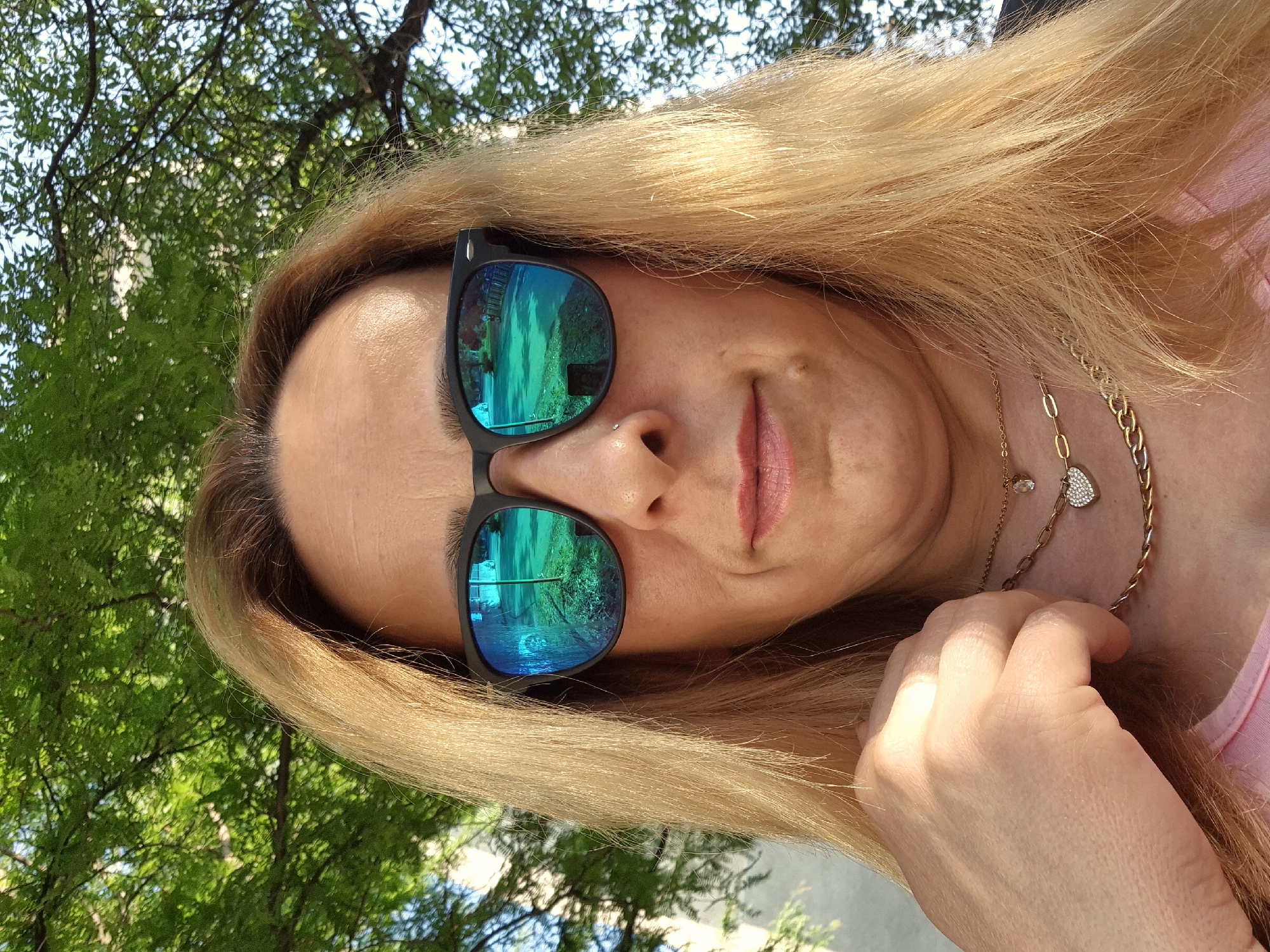 A close-up, eye-level portrait shows an adult individual with long, straight, blonde hair with visible darker roots, wearing black-framed sunglasses with reflective blue-green lenses, outdoors in the city of Unknown, Unknown. The individual's face is framed by the hair, which extends down past the shoulders. A small silver stud piercing is visible on the individual's right nostril. Below the individual's chin, a small, dark mole is present. Multiple gold-toned necklaces adorn the neck: a thin chain with a small clear stone pendant, another thin chain with a heart-shaped pendant encrusted with small clear stones, and a thicker curb chain. A light pink garment is visible on the upper torso. The individual's left hand, with fingers slightly curled, is positioned near the lower jawline/upper chest. The reflective lenses of the sunglasses show a distorted mirror image of an outdoor scene, including green foliage, a paved area, and a vertical green pole; the right lens reflection also shows faint outlines of distant figures and a fence-like structure. The background behind the individual consists of out-of-focus green tree leaves under natural sunlight, with hints of a light blue sky.