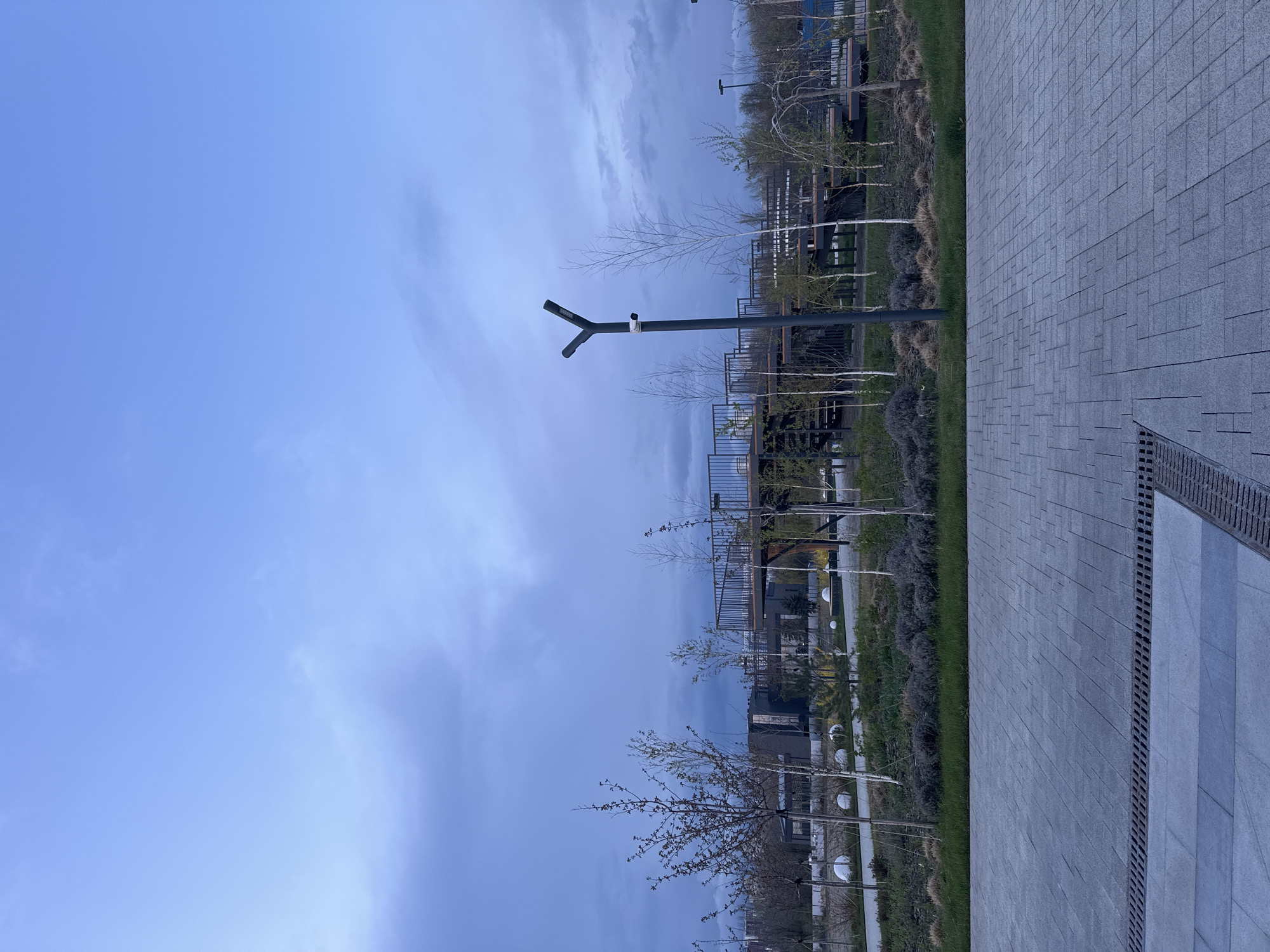 The image shows a park in Rostov-on-Don, Russia. A grey brick path runs along the right side of the frame. Next to the path is a narrow strip of grass and small trees. A black lamppost stands near the center of the image. In the background, there are modern buildings and more trees. The sky is a pale blue color with streaks of clouds.