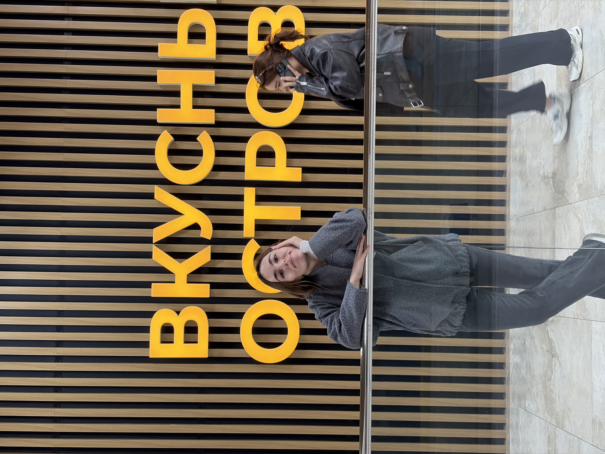 The image shows a scene in Izhevsk, Russia, featuring a reflective surface, likely a storefront window. On the left, a textured wall is visible, with horizontal wooden slats, some light brown, others darker. Overlaid on the wall are large, vertically arranged yellow Cyrillic letters spelling out "ВКУСНО – И ТОЧКА" (Vkusno — i tochka), a Russian fast food chain.
Two people are reflected in the window. On the top, a woman wearing a dark leather jacket and dark pants is holding a phone to her ear. Her hair is pulled back in a ponytail. Her reflection shows her from the waist down.
The second reflection depicts a woman wearing a gray sweater and dark jeans. She is leaning on the window, her face visible. Her reflection also shows her from the waist down. The ground is visible and appears to be light-colored tile.