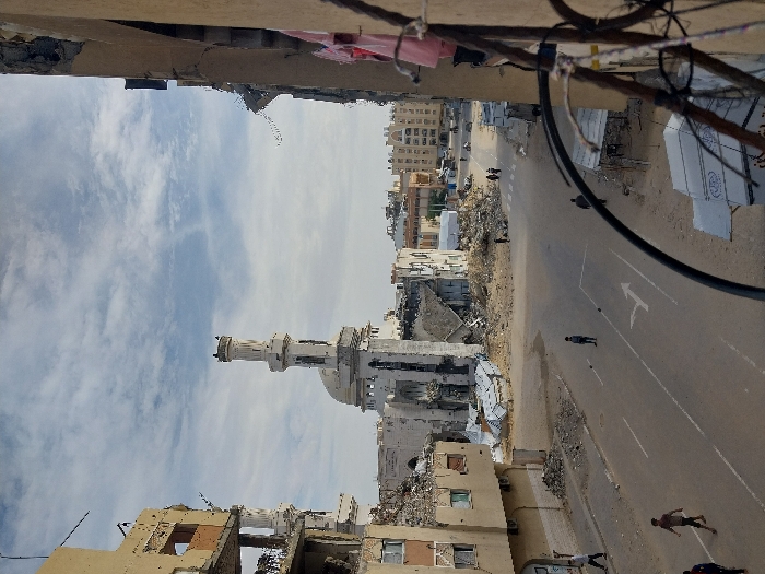 This image depicts a wide-angle view of a street scene in Khan Yunis, Palestinian Territory, characterized by significant urban destruction under a partly cloudy sky. In the foreground, the frame is partially obscured on the right by a dark, curved cable or wire.
To the left, a multi-story building stands heavily damaged, with large sections of its facade collapsed, exposing internal structures, rebar, and debris. Multiple windows are either missing or shattered. Opposite this, on the far right, another building's damaged exterior is visible, with articles of clothing (including a pink item) hanging from a railing or window.
Dominating the central background is a large mosque with a prominent white dome and an tall, ornate minaret, also exhibiting visible damage and surrounded by extensive rubble at its base. Behind the mosque, further buildings, some appearing intact and others damaged, line the horizon.
The asphalt street in the mid-ground runs from left to right, marked with faded white lines, including a right-pointing arrow. The areas immediately adjacent to the damaged buildings on both sides of the street are covered in piles of concrete debris, twisted metal, and general rubble.
Several individuals are present on the street. In the mid-ground, three men walk on the asphalt: one on the far left walking away from the viewer, another in the center walking towards the viewer, and a third to the right walking away. In the distance, a group of approximately four individuals walks on the right side of the road. On the right side of the street, closer to the foreground, there are what appear to be makeshift structures or debris piles.