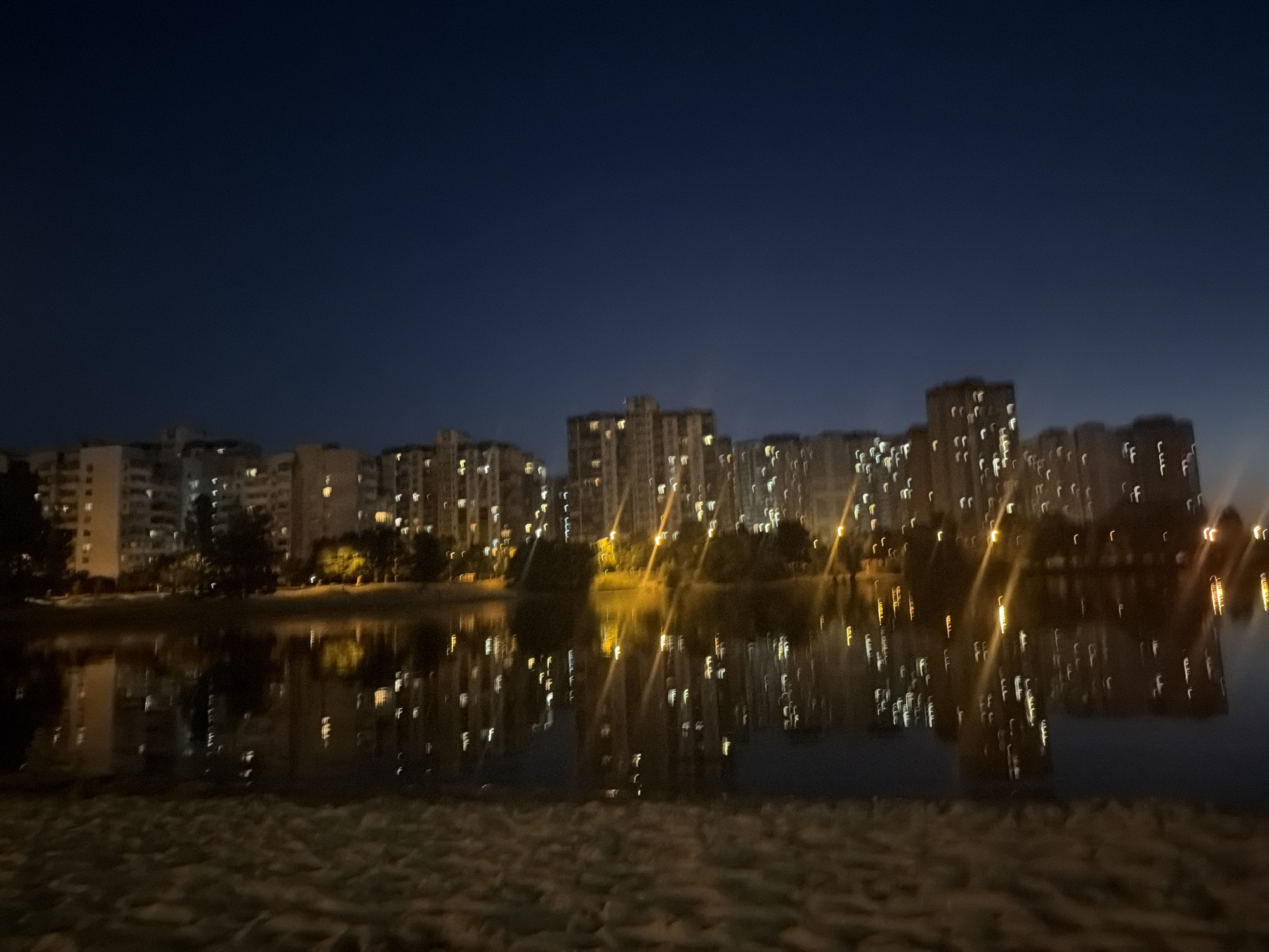 The media file depicts a nighttime scene of a city skyline reflected in a body of water. The image is characterized by a dark, clear night sky, with a deep blue hue indicating the absence of clouds and the fading light of dusk or the early hours of dawn.
The primary subjects are a series of tall apartment buildings, illuminated by numerous windows, suggesting a residential area. These buildings are arranged in a row along the far bank of a body of water, likely a river or a lake. The lights from the buildings are sharply reflected in the water, creating a shimmering, almost abstract pattern of light and dark.
The foreground shows a sandy or gravelly area, possibly a beach or a pathway, adding a textural element to the scene. Trees and vegetation are visible along the waterfront, interspersed with the buildings, adding a touch of nature to the urban landscape.
The overall impression is one of tranquility and urban beauty at night. The reflections in the water are particularly striking, creating a sense of depth and mirroring the city's verticality. The time of day is clearly nighttime, as indicated by the dark sky and the illuminated windows. There is no visible text in the image. The location context provided is Kyiv, Ukraine.