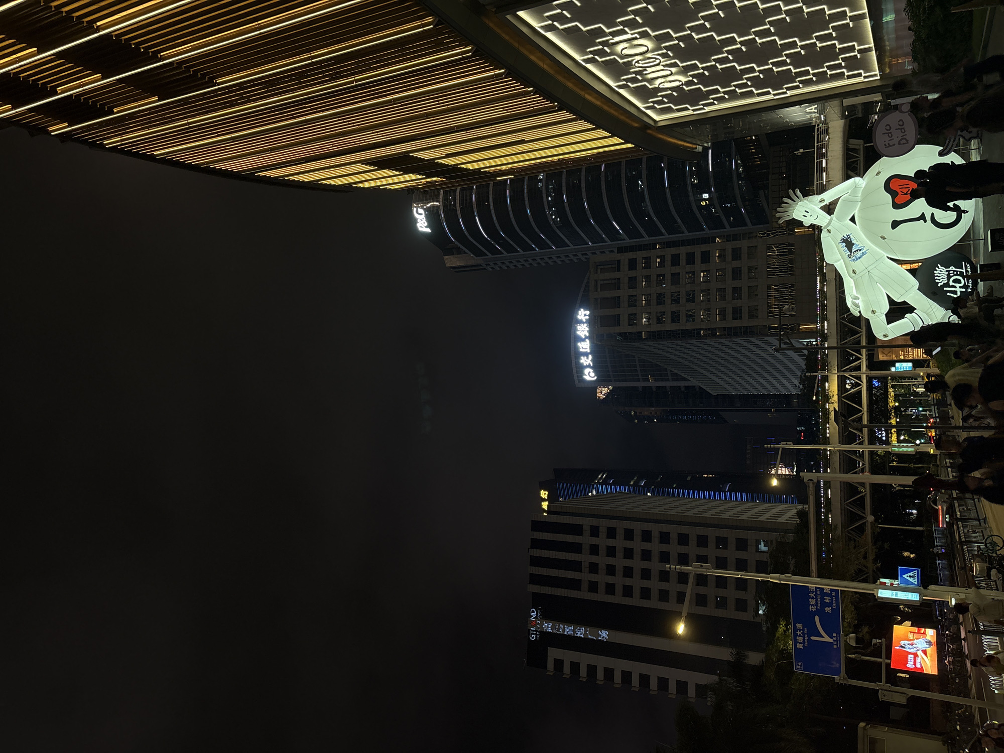 The image depicts a nighttime scene in Tianhe District, China, characterized by towering, illuminated skyscrapers and a bustling street. In the foreground, a large, inflatable sculpture of a stylized figure rests on a spherical object bearing the text "I ♥ Kili" and "Fido Dido." Several individuals are visible, some interacting with the inflatable, while others are walking or standing along the street. A prominent building on the right features the "GUCCI" logo illuminated in a grid pattern. Other buildings in the background display signage for "P&G" and "GILAND." Streetlights and traffic lights illuminate the roadway, and a pedestrian overpass is visible in the mid-ground. The overall atmosphere is urban and lively, with the city's architecture and lighting creating a vibrant nocturnal landscape.