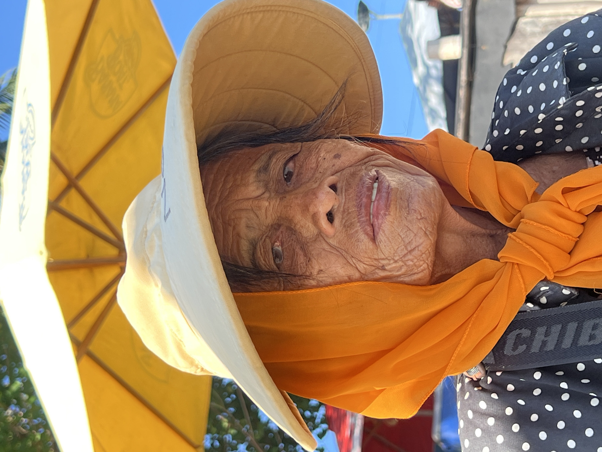 The image shows a close-up of an elderly woman wearing a wide-brimmed yellow hat and an orange scarf. She is also wearing a black and white polka dot top. Her face is deeply wrinkled, suggesting an older age. The woman is looking towards the camera with a neutral expression.
Behind her, a yellow umbrella is visible, with some text on it, although it is partially obscured and difficult to read. The umbrella and the clear blue sky suggest an outdoor setting, likely during the daytime. The overall lighting indicates it's a sunny day.
The woman's attire, particularly the scarf and polka dot top, along with the presence of a beach umbrella, hints at a tropical or coastal location. Given the location context of Kuta, Indonesia, it is probable she is at a beach or a similar outdoor public space. The image captures a moment of candid portraiture, focusing on the woman's face and her immediate surroundings.