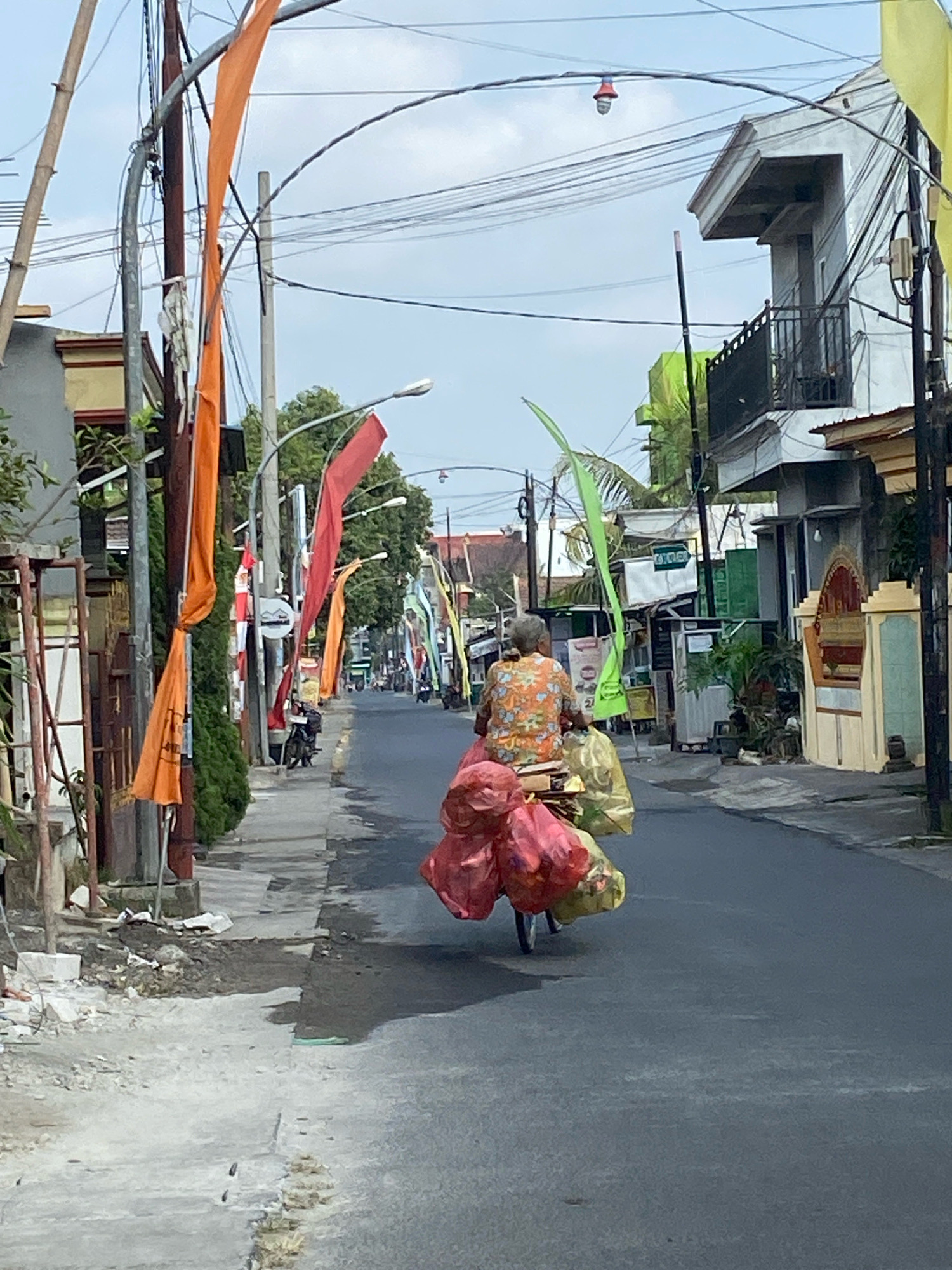 The image depicts a street scene in Kediri City, Indonesia, characterized by a person riding a bicycle laden with goods. The rider, seen from behind, is wearing a colorful, patterned shirt and is seated on the bicycle, which is equipped with a rear rack carrying an assortment of items, predominantly red and pink plastic bags, along with some yellow bags, likely filled with produce or other merchandise. The bicycle also appears to have a basket or carrier in front of the rider.
The environment is an urban street with buildings lining both sides. The road surface is asphalt, and a concrete sidewalk is visible to the left, showing signs of disrepair and construction or debris. Overhead, a tangle of electrical wires and streetlights adorn the scene. Colorful flags, predominantly orange and red, are strung along the street, suggesting a festive occasion or local decoration. The buildings are varied in style, some appearing to be residential homes with simple architecture, while others are more complex, multi-story structures. Vegetation, including trees and bushes, is interspersed throughout the urban landscape. The sky is overcast, contributing to the diffused lighting conditions of the image. Several other individuals and vehicles, including motorcycles, are present in the background, indicating a moderate level of activity on the street.
