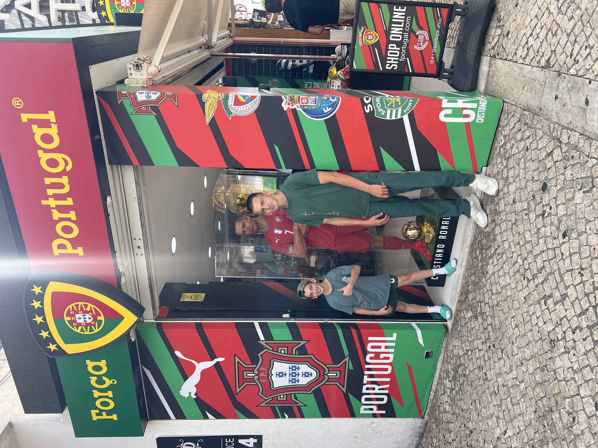 The image shows the exterior of a shop in Albufeira, Portugal, specializing in Portuguese football merchandise. The shop's facade features the "Força Portugal" slogan and a crest, with the Portuguese national colors of red and green prominently displayed in a diagonal stripe pattern. A life-sized Cristiano Ronaldo mannequin is visible inside the shop, alongside football trophies. Two young men stand outside the shop, one in the foreground giving a thumbs-up. One is wearing a green t-shirt and shorts, and the other is wearing a green t-shirt and pants. The shop entrance displays a "SHOP ONLINE fportugal.com" sign, along with social media icons. The street is paved with traditional Portuguese cobblestones.