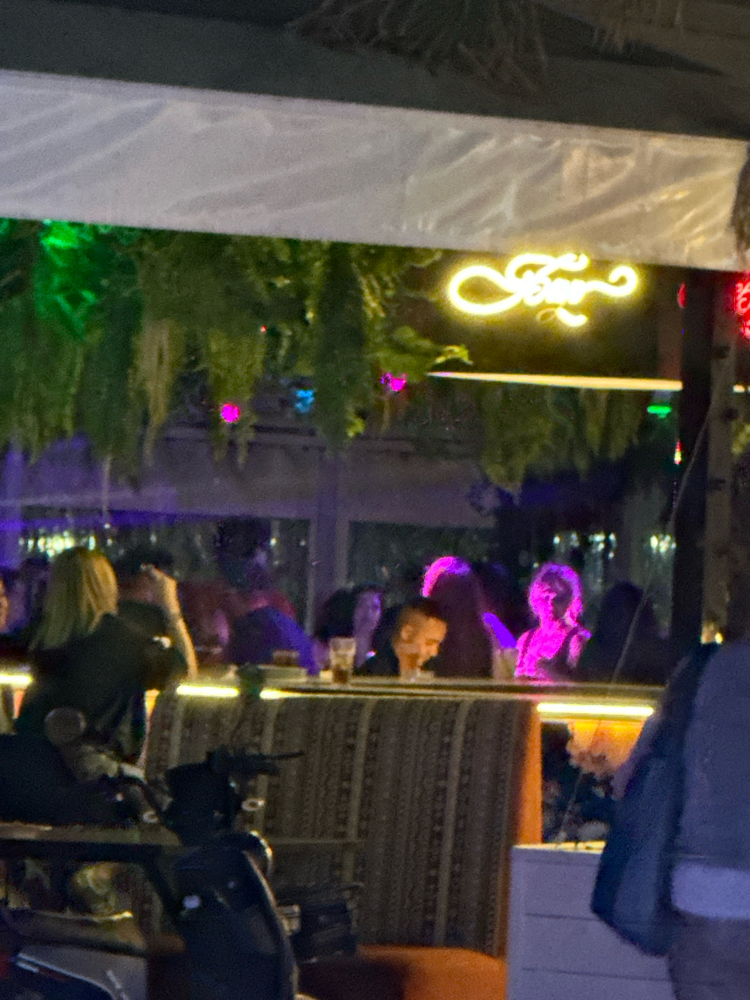 An outdoor seating area at a venue in Lermontovo, Russia, is depicted at night. A neon sign with stylized script in a bright yellow hue is visible overhead, emitting a warm glow. Green foliage hangs down from above, interspersed with small, colorful lights that cast purple and blue hues. Several individuals are seated at tables, some appearing engaged in conversation. A motorcycle is parked in the foreground, partially obscuring the seating area. The overall atmosphere suggests a lively evening gathering.