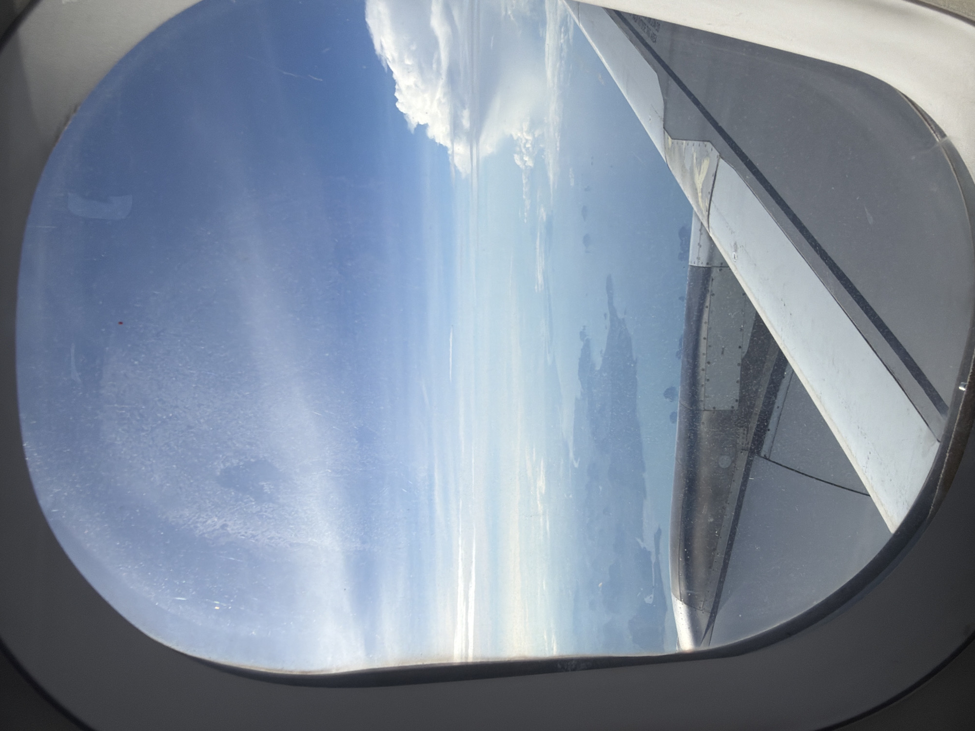 The image, taken from an airplane window, offers a view of a coastal landscape, likely near Ko Yao Noi, Thailand. The foreground is dominated by the wing of the airplane, with its metallic engine housing and a section of the wing itself visible. Beyond the wing, the turquoise sea stretches out, dotted with numerous small islands and landmasses that appear to be part of Ko Yao Noi. The islands are covered in lush greenery and are surrounded by the calm water. In the distance, the sky is a brilliant blue, with scattered white clouds, including a large cumulonimbus cloud formation on the right side of the frame. The sunlight creates a bright haze, suggesting it is daytime. The window itself is slightly smudged, with a few streaks and particles visible on the glass.