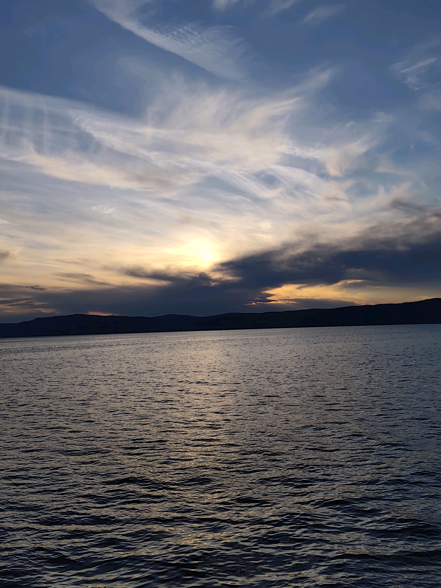 The image depicts a tranquil, serene body of water, likely Lake Baikal, at dusk, with the distant silhouette of mountains along the horizon. The sky is a dramatic canvas of blues and grays, streaked with wispy white clouds that catch the fading sunlight. The sun itself is partially obscured by dark clouds, casting a warm, golden glow on the water's surface, creating a shimmering effect of light and shadow. The water is calm, with gentle ripples suggesting a peaceful atmosphere. The scene evokes a sense of vastness and natural beauty. Given the geographical context of Listvyanka, Russia, which is situated on the shores of Lake Baikal, this landscape is characteristic of the region, particularly during sunset. There are no people or discernible man-made structures visible in the image, emphasizing the untouched natural state of the environment.