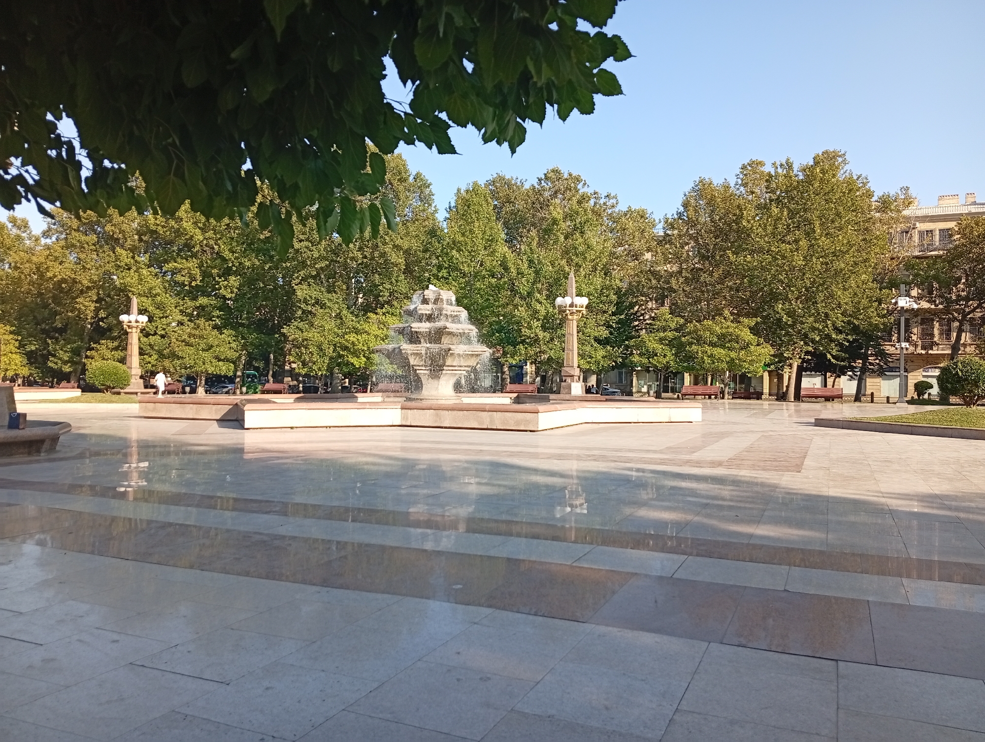 The image depicts a sunny day in Baku, Azerbaijan, specifically in the Fountain Square area. The foreground is dominated by a polished, light-colored tiled plaza, wet in places and reflecting the sky and surrounding structures. A set of shallow steps leads up from the foreground plaza to a raised section. In the mid-ground, a multi-tiered fountain is active, with water cascading down its levels. Flanking the fountain are ornate lamp posts. Behind the fountain, a line of lush green trees forms a backdrop, with some buildings visible beyond them. The buildings appear to be in the European architectural style common to Baku, with visible windows, balconies, and chimneys. A few red benches are visible beneath the trees, and a solitary figure in white is walking in the distance on the raised plaza. The overall scene is one of a well-maintained public space on a clear day.