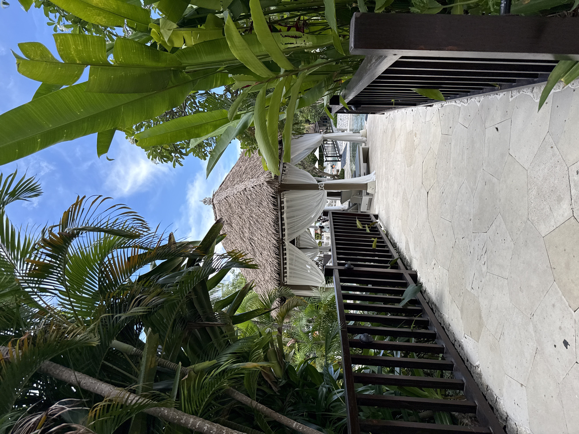 The image captures an outdoor scene in Nusa Dua, Indonesia, featuring a pathway leading towards a thatched-roof structure adorned with white curtains. The pathway is constructed from irregularly shaped stone tiles and is bordered by a dark wood railing on one side. Lush tropical foliage, including palm trees and banana leaves, frames the scene. The sky is visible through the foliage, showing a blue expanse with scattered clouds. A set of wooden stairs runs alongside the pathway, close to the wooden railing. The overall atmosphere is tropical and serene.
