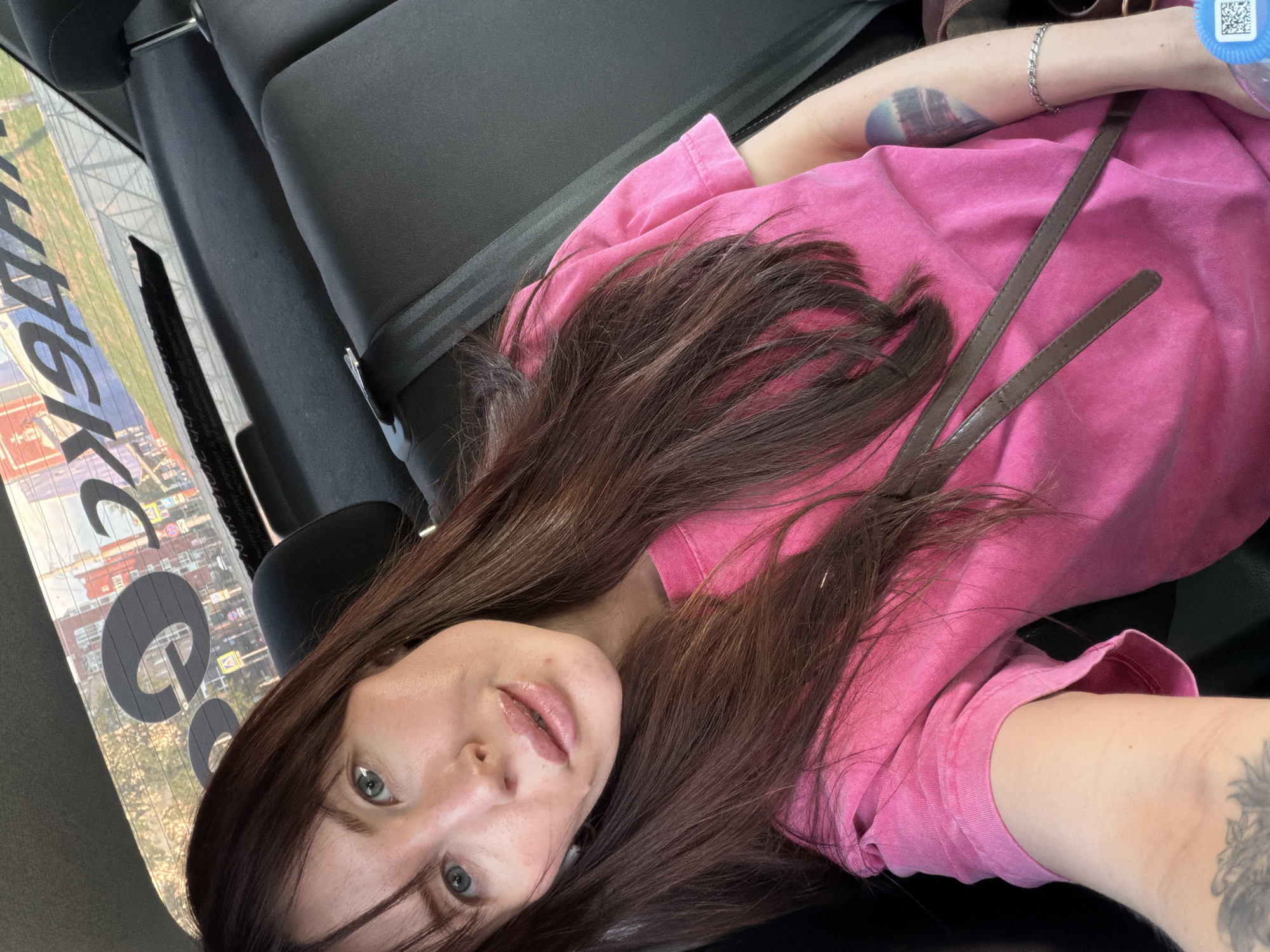 The image shows a woman in the backseat of a vehicle, wearing a pink t-shirt and seatbelt. She has long brown hair and light-colored eyes. A purse strap is visible over her shoulder, and tattoos are present on her arms. Through the car window, a cityscape is visible with signage indicating the location is Perm, Russia.