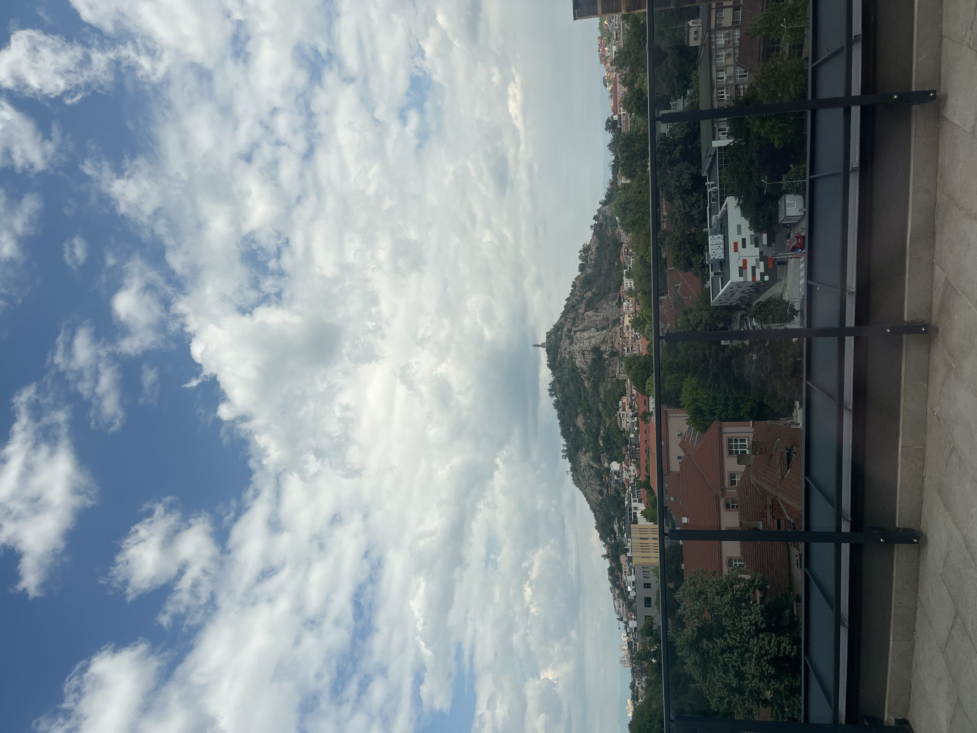 The image shows a view from a high vantage point, likely a balcony, overlooking a cityscape in Plovdiv, Bulgaria. In the distance is a prominent hill known as Bunardzhik Hill, topped by the Alyosha Monument (a statue of a Soviet soldier). The hill is surrounded by buildings of varying heights and architectural styles, with red-tiled roofs predominating. Lush greenery, including trees and shrubs, dots the landscape. The sky is filled with a mix of blue patches and white clouds. The foreground features a metal railing and a tiled surface, suggesting the image was taken from a balcony. No people are visible.