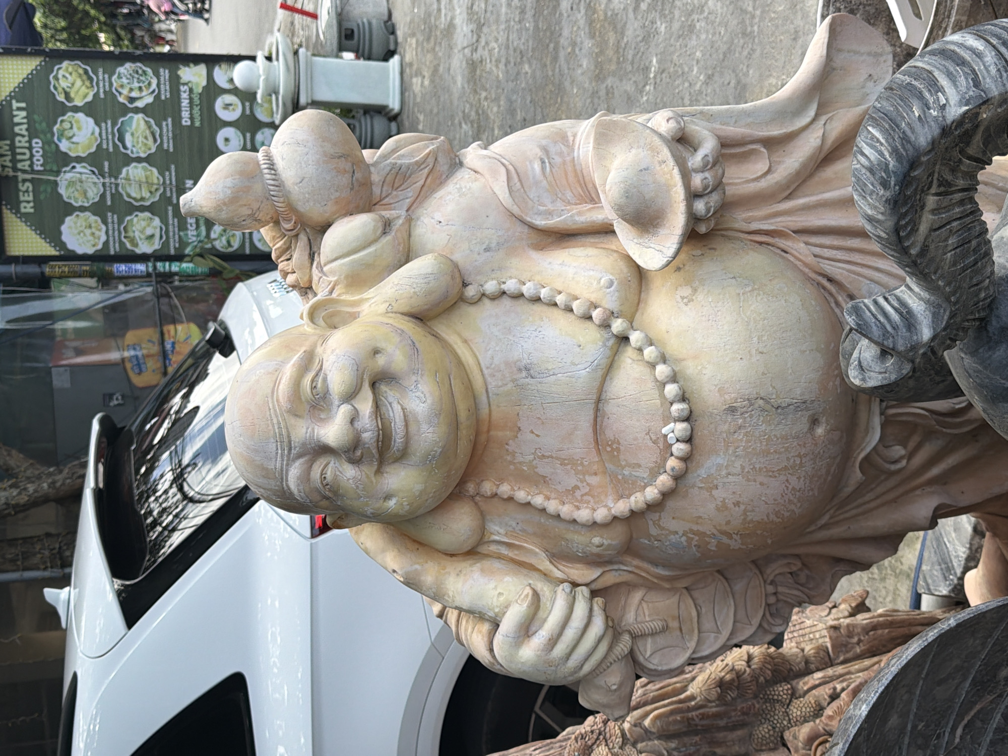 A life-sized, tan-colored statue of a laughing Buddha is displayed outdoors, likely in a market or shop specializing in garden ornaments or religious artifacts. The Buddha statue is intricately detailed, with a round face, large belly, and a necklace of round beads. It is holding a gold ingot or bowl in its right hand and a gourd-shaped object on its left shoulder. The statue has a weathered appearance, with visible texture and coloration that mimics aged stone or wood.
In the background, a white SUV is partially visible on the left, with a car antenna on its roof. To the right of the statue, a green menu board for "SAM RESTAURANT FOOD" is displayed, featuring images of various dishes and drinks. A light-colored decorative pillar is also present to the right of the Buddha. The ground is paved with concrete or stone tiles, suggesting an urban outdoor setting.
The scene appears to be located in Đà Nẵng, Vietnam. This is inferred from the presence of the Vietnamese restaurant name and the general aesthetic of outdoor market stalls often found in Vietnamese cities. The presence of people in the background, though blurred, indicates a public space. The overall atmosphere is that of a typical commercial street scene in Vietnam, showcasing local culture and commerce.
