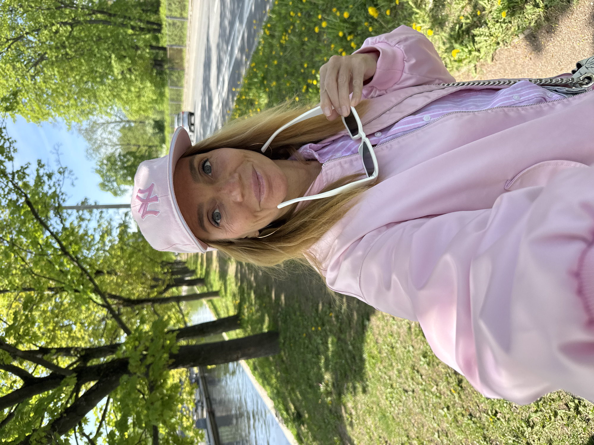 A woman wearing a pink baseball cap, a pink jacket over a pink and white striped shirt, and gold hoop earrings is holding a pair of white sunglasses. She is standing outdoors on a patch of grass near a canal, with trees lining the bank. In the background, there is a road, and yellow dandelions dot the landscape. This is likely in Saint Petersburg, Russia, given the location and architecture.