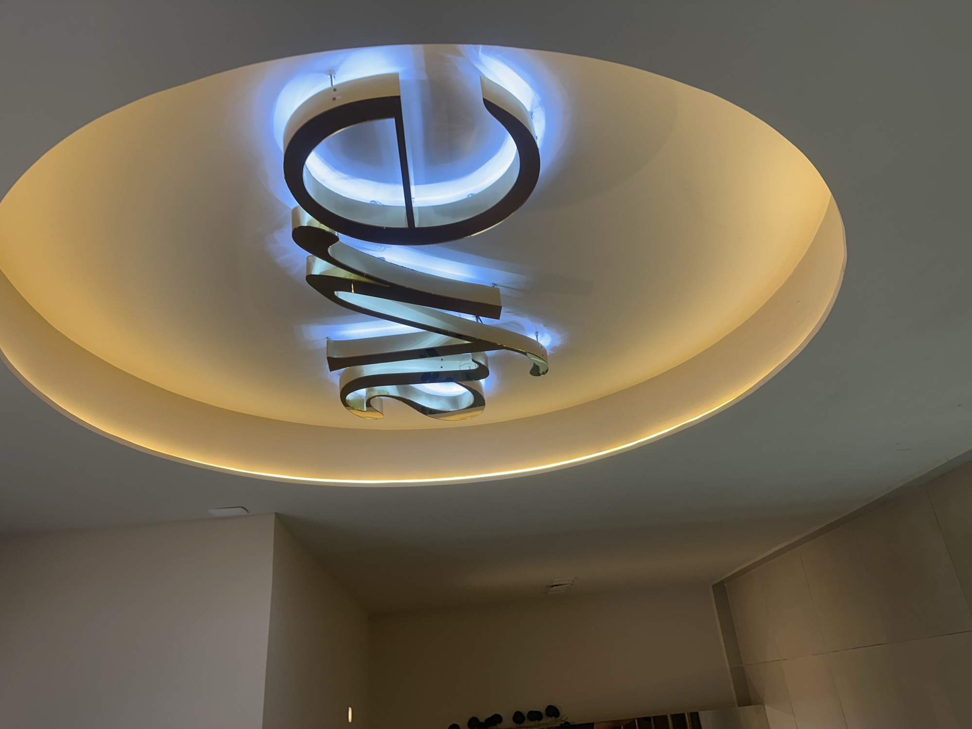 The image depicts an interior view of a room in San Bernardino Tlaxcalancingo, Mexico. The primary subject is a large, circular, recessed ceiling fixture. The fixture is layered, with a central design that appears to spell out "ONE" in a stylized, vertically stacked, illuminated manner. The lettering is rendered in a combination of dark and light materials, potentially metals or painted elements, with a contrasting blue and white glow emanating from behind the letters. The overall shape of the fixture is circular, surrounded by a warm, ambient light along the edge of the recessed portion of the ceiling.
The room itself features light-colored walls. The lower portion of the image includes glimpses of interior elements, such as a neutral-colored wall on the left and a section of wall with vertical panels on the right. There are indistinct objects on the floor beneath the ceiling fixture. The image lacks clear human presence, focusing instead on the architectural details and lighting.