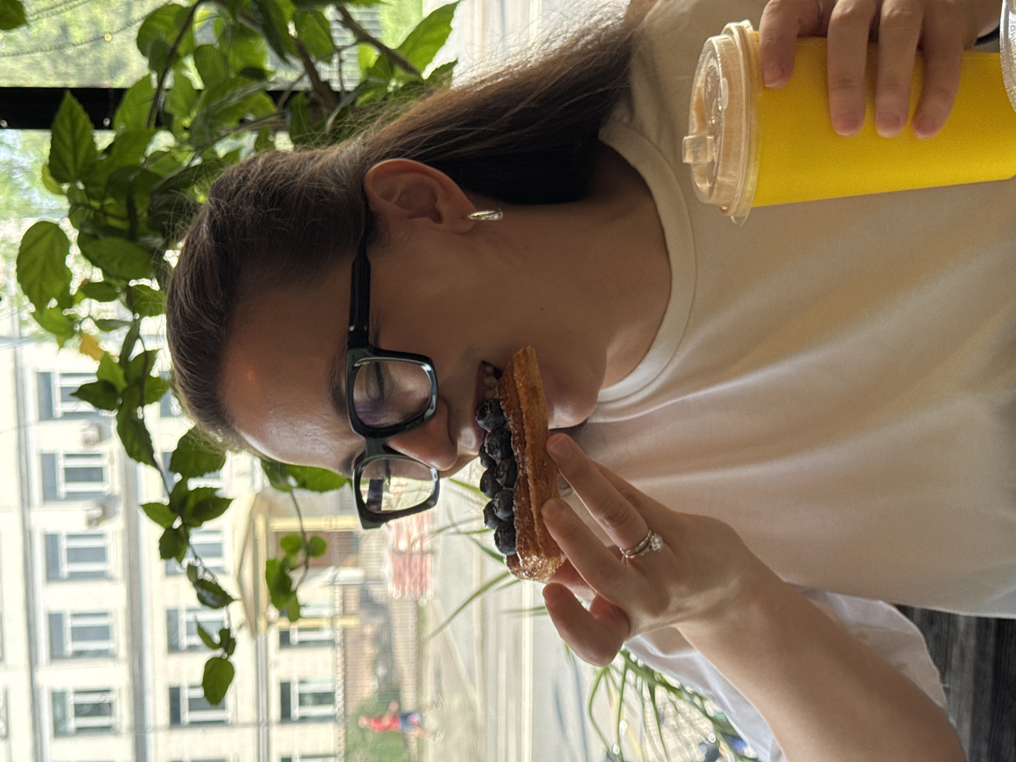 A woman with long brown hair and glasses is eating a dessert topped with blueberries while holding a yellow drink in her hand. The dessert appears to be some type of pastry, possibly a churro. She is wearing a white shirt and a ring on her finger. Through the window behind her, a building with multiple windows can be seen. Based on the architectural style of the building and surrounding foliage, the location is possibly in Moscow, Russia.