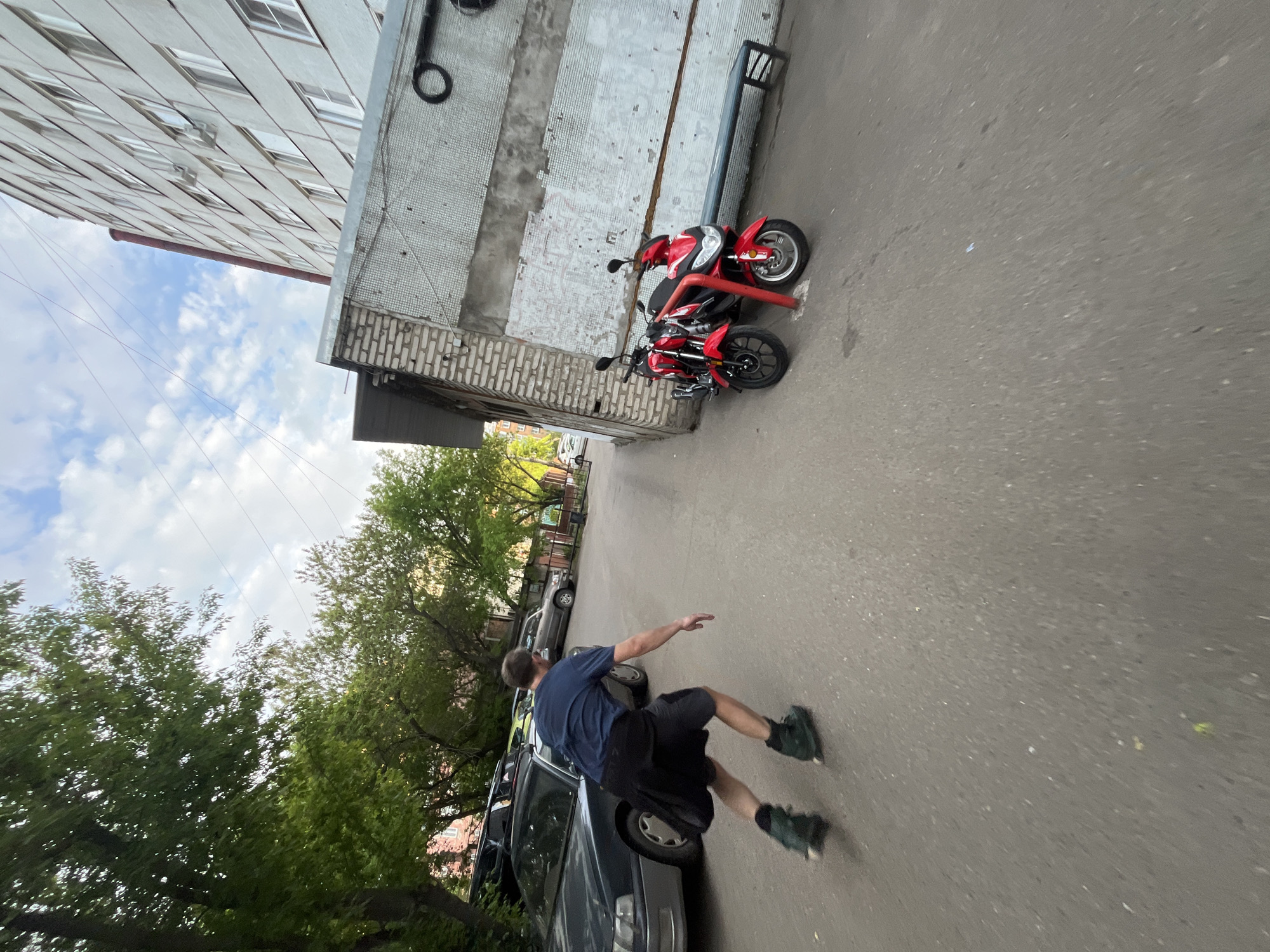 A photograph taken in Krasnoyarsk, Russia shows a man walking on a paved road near a building. The man is wearing a dark blue shirt and dark shorts with dark green shoes. He has his right arm extended. To the left is a parked dark grey car and trees. To the right are two red motorcycles parked near a white building. In the background are more trees and buildings with a partly cloudy sky visible above.