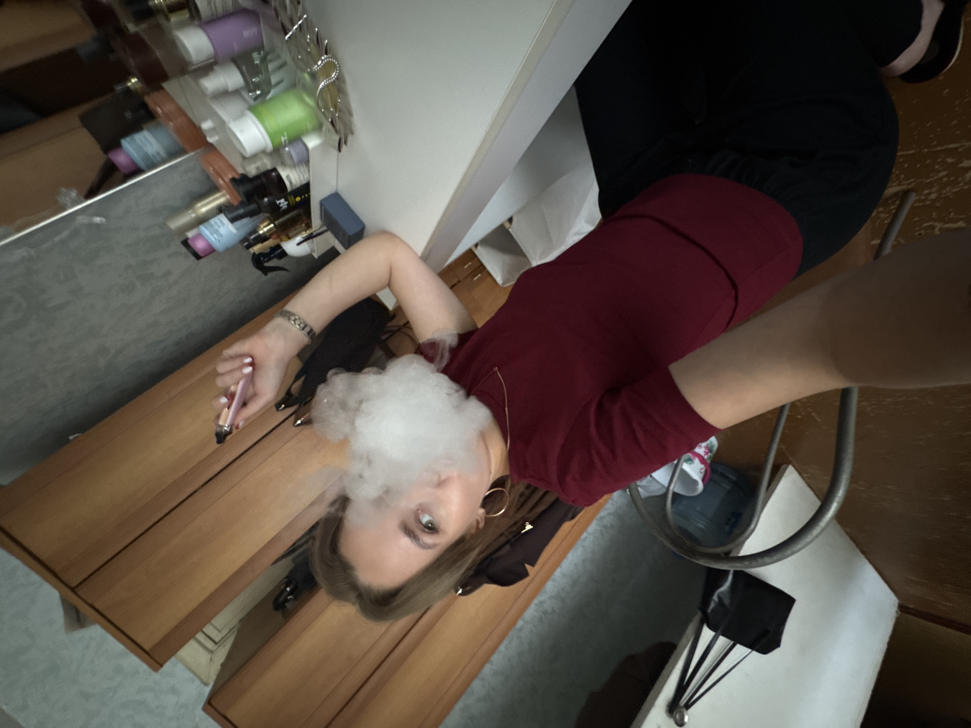 The image shows a young woman with blonde hair, wearing a maroon long-sleeved shirt and black pants. She is holding a pink vape pen in her right hand and exhaling a cloud of vapor. The photo appears to be a selfie taken in a room, possibly a bedroom or bathroom, with wooden furniture and shelves filled with various items, including bottles and what look like beauty products.
The lighting suggests it might be daytime. The woman's expression is neutral, and the overall mood is casual. The background includes a mirror reflecting more shelves and products, a white wall, and some darker elements that are difficult to identify clearly. The presence of a vape pen indicates the activity of vaping. The scene is indoors, with domestic elements visible. There is no discernible text in the image.