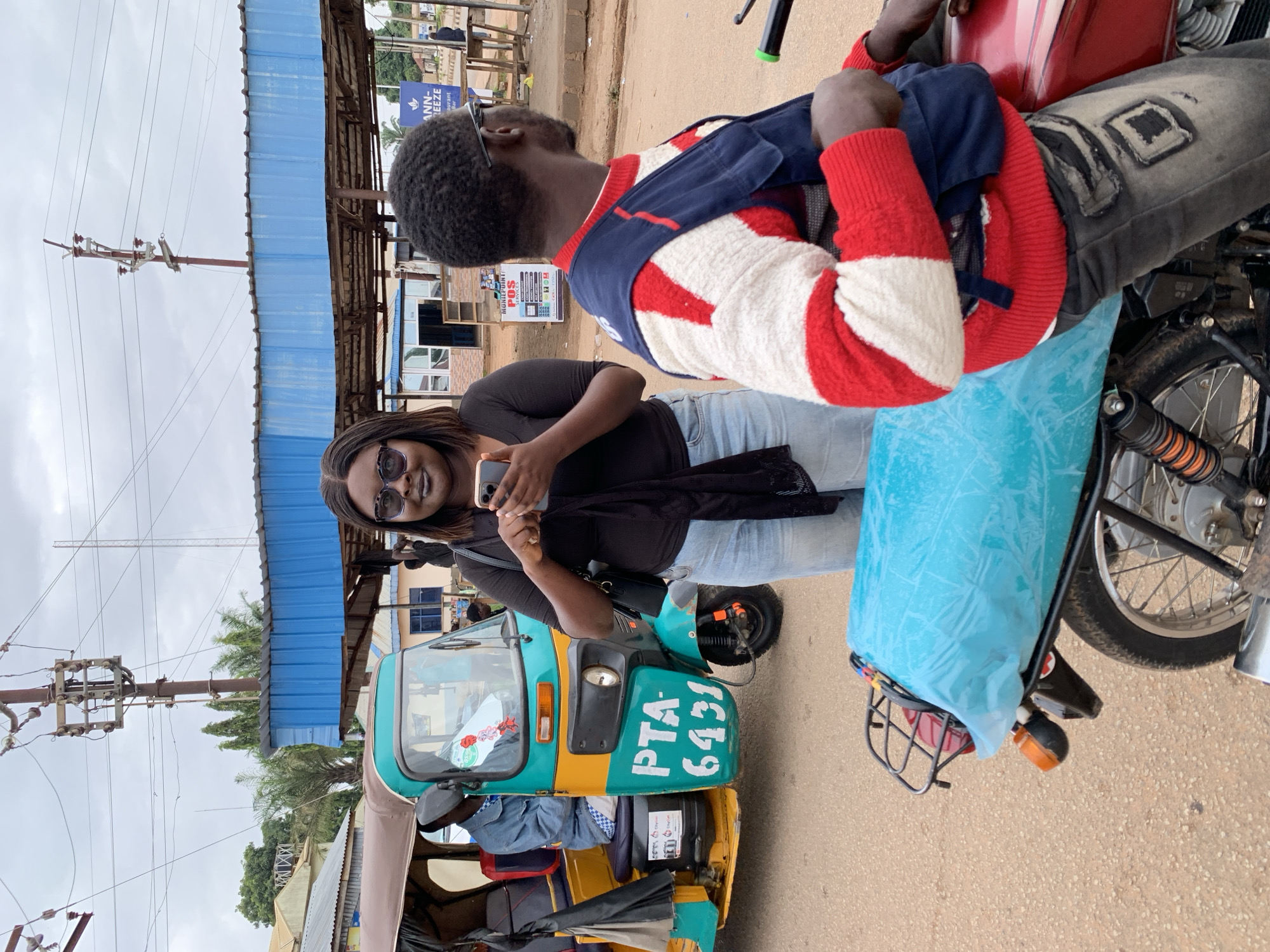 In Gura Top, Nigeria, a medium shot captures a street scene with a woman and two men. The woman, positioned centrally and facing the camera, wears sunglasses and a black top with a black scarf draped over her shoulders. She is holding a smartphone in her right hand, appearing to be using it. To her left, a three-wheeled auto-rickshaw, predominantly teal and yellow with "PTA 6431" visible on its side, is parked. A man in a blue jacket is seated in the driver's position of the auto-rickshaw. To the right of the woman, a man wearing a red and white striped sweater and a dark blue vest is seated on a red motorcycle. His back is partially to the camera. The environment suggests an outdoor, possibly commercial area, with a blue awning structure and buildings visible in the background. Power lines and utility poles are present overhead, indicating an urban or semi-urban setting. The ground is paved with a sandy, reddish-brown material, typical of roads in the region.
