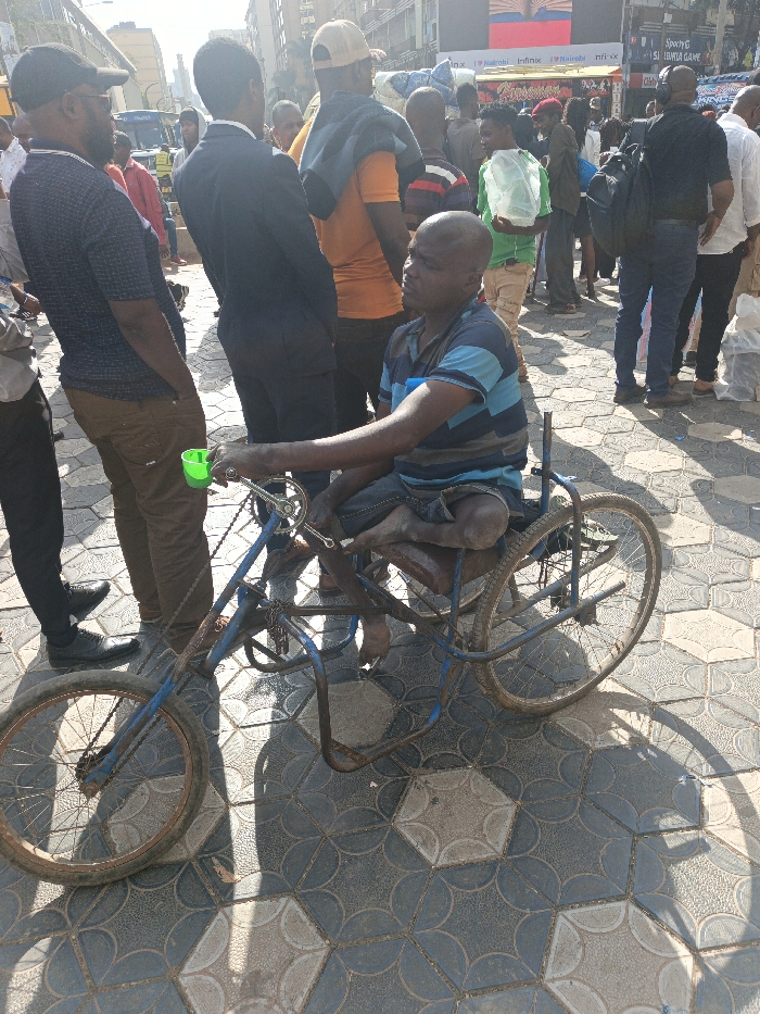 The image depicts a crowded outdoor urban scene in Nairobi, Kenya, under bright sunlight. In the foreground, a bald, dark-skinned adult male, wearing a blue and light blue horizontally striped short-sleeved shirt and dark shorts, is seated on a custom-built, blue-painted three-wheeled mobility device. His legs appear significantly shortened or amputated below the knees, with his bare feet resting on a platform. He operates the device via hand cranks, one of which has a small green plastic cup attached. He is looking slightly to the right with a neutral expression.
To the immediate left of the central figure stands another dark-skinned adult male, wearing a dark patterned short-sleeved shirt, brown trousers, a dark baseball cap, and sunglasses. His hands are in his pockets, and he is looking forward. Behind him and slightly to the left, a third dark-skinned adult male, dressed in a dark suit jacket and trousers, stands facing away from the camera, looking to the right.
The background is densely populated with a diverse crowd of predominantly dark-skinned adults, mostly male, engaged in various activities such as walking and standing. They wear a range of casual clothing. Notable individuals include a man in an orange t-shirt with a beige cap and a blue garment draped over his shoulder, and another in a green shirt holding a clear plastic container.
The ground is paved with interlocking grey and dark grey hexagonal tiles. Strong shadows are cast on the pavement, indicating intense sunlight from the upper left. In the far background, multi-story urban buildings are visible. One prominent building displays a large digital billboard with text including "I love Nairobi" and "Infinix," definitively placing the location in Nairobi, Kenya. A blue bus is partially visible on a street in the distant left background.