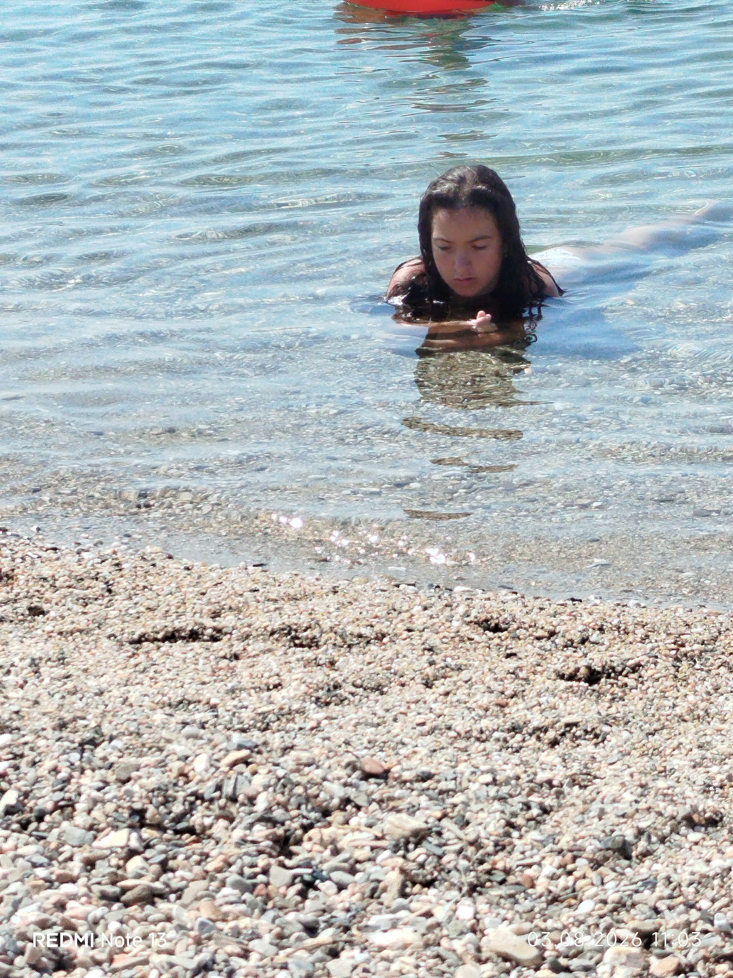 A young girl with dark hair is swimming in clear, shallow water with a pebble beach in the foreground. She is looking down at her hands, which are clasped together. Her body is mostly submerged in the water, and a faint outline of what appears to be a mermaid tail is visible behind her. The water is calm with gentle ripples, and sunlight is reflecting off its surface. The beach is covered in smooth, multicolored pebbles. In the upper portion of the image, a blurred red object, possibly a float or inflatable, is visible in the water. A timestamp in the lower right corner reads "03.08.2026 11:03".