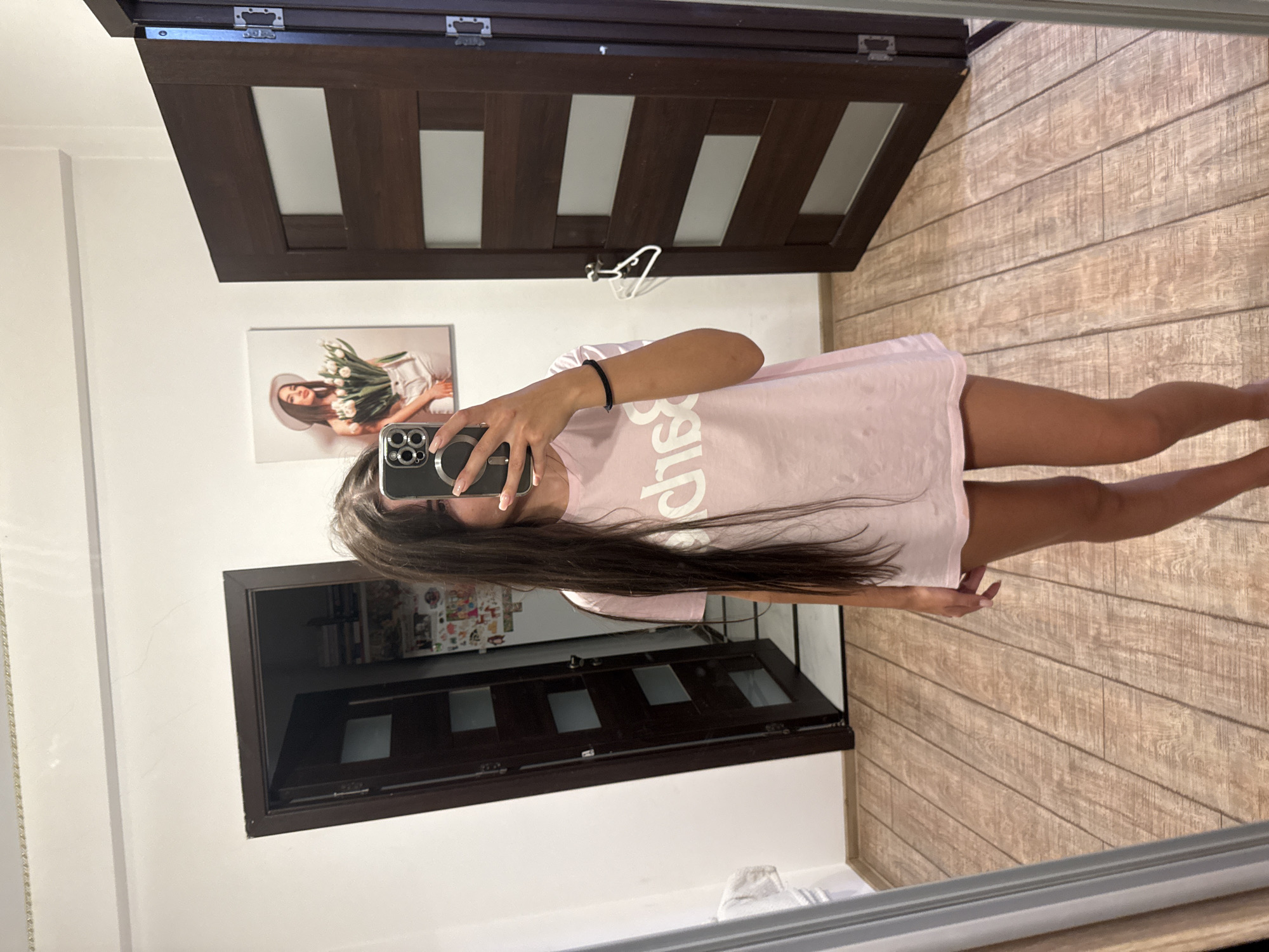 In a domestic interior, a young woman with long, dark hair stands in front of a mirror, holding a smartphone in her right hand to take a selfie. She is wearing a light pink t-shirt with "Baci" printed on it in white letters. Her left arm is slightly bent, with a black bracelet on her wrist. The background reveals a hallway with two dark wooden doors. One door on the left is ajar, showing a glimpse of a cluttered room with shelves. The door on the right is closed, with a white hanger hooked onto its handle. A framed picture of a woman holding a bouquet of tulips hangs on the wall between the two doors. The floor is covered with light brown wooden planks. The scene is consistent with a residential setting, potentially in Chișinău, Moldova, given the provided context, however, there are no specific visual cues within the image to confirm this location.
