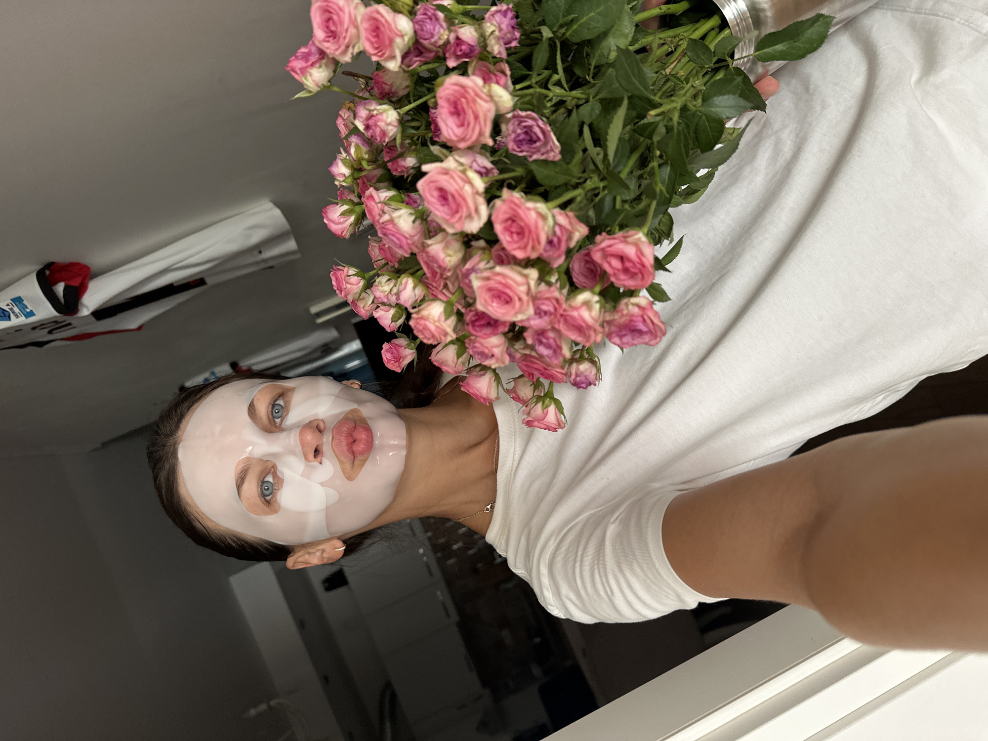 A young woman with blue eyes and a sheet mask on her face is holding a bouquet of pink roses in a silver vase. She is wearing a white t-shirt and a gold necklace. The background is a dimly lit room with a wall and a doorframe. There is also a glimpse of a jersey hanging on the wall in the background. The scene is likely indoors, possibly in a residential setting in Boreti, Montenegro.