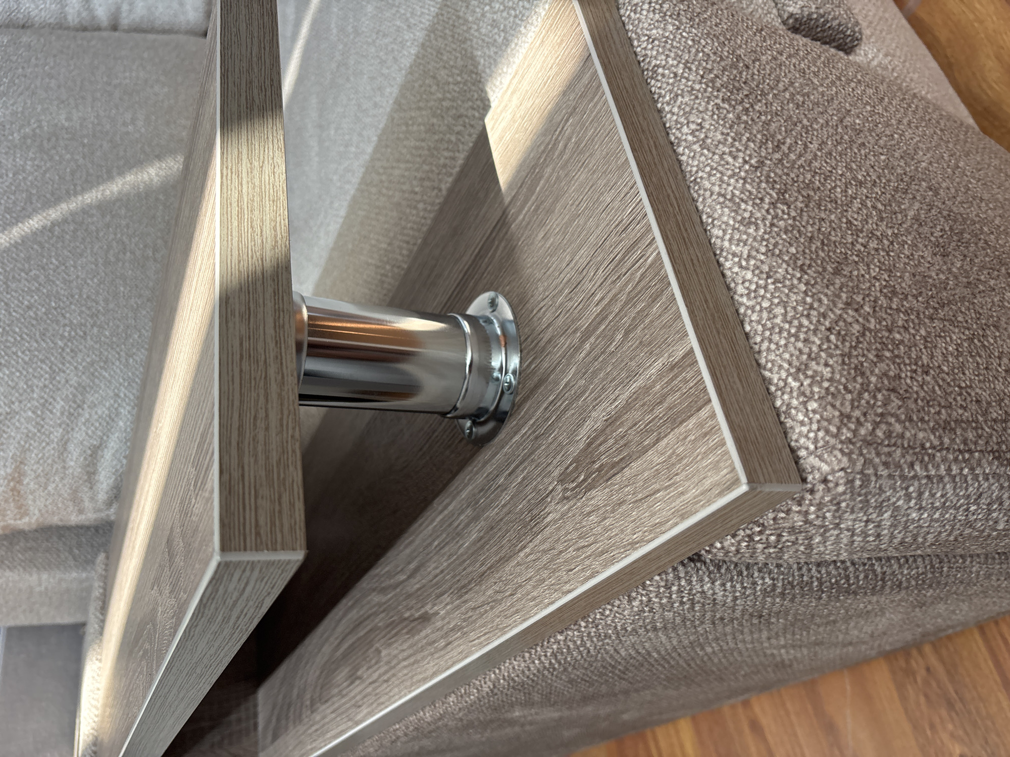 This is a close-up, high-angle shot of a metallic cylinder attached to a wooden shelf. The cylinder is polished and reflects light. The shelf has a wood-grain pattern and a light-colored edge. The background is out of focus, showing a textured, neutral-colored fabric, likely part of a sofa or upholstered furniture. Sunlight is casting shadows and highlights across the scene, suggesting it is daytime and possibly near a window. There is no visible text. The scene appears to be indoors, with hints of a wooden floor visible at the bottom right. The setting is likely a domestic environment.