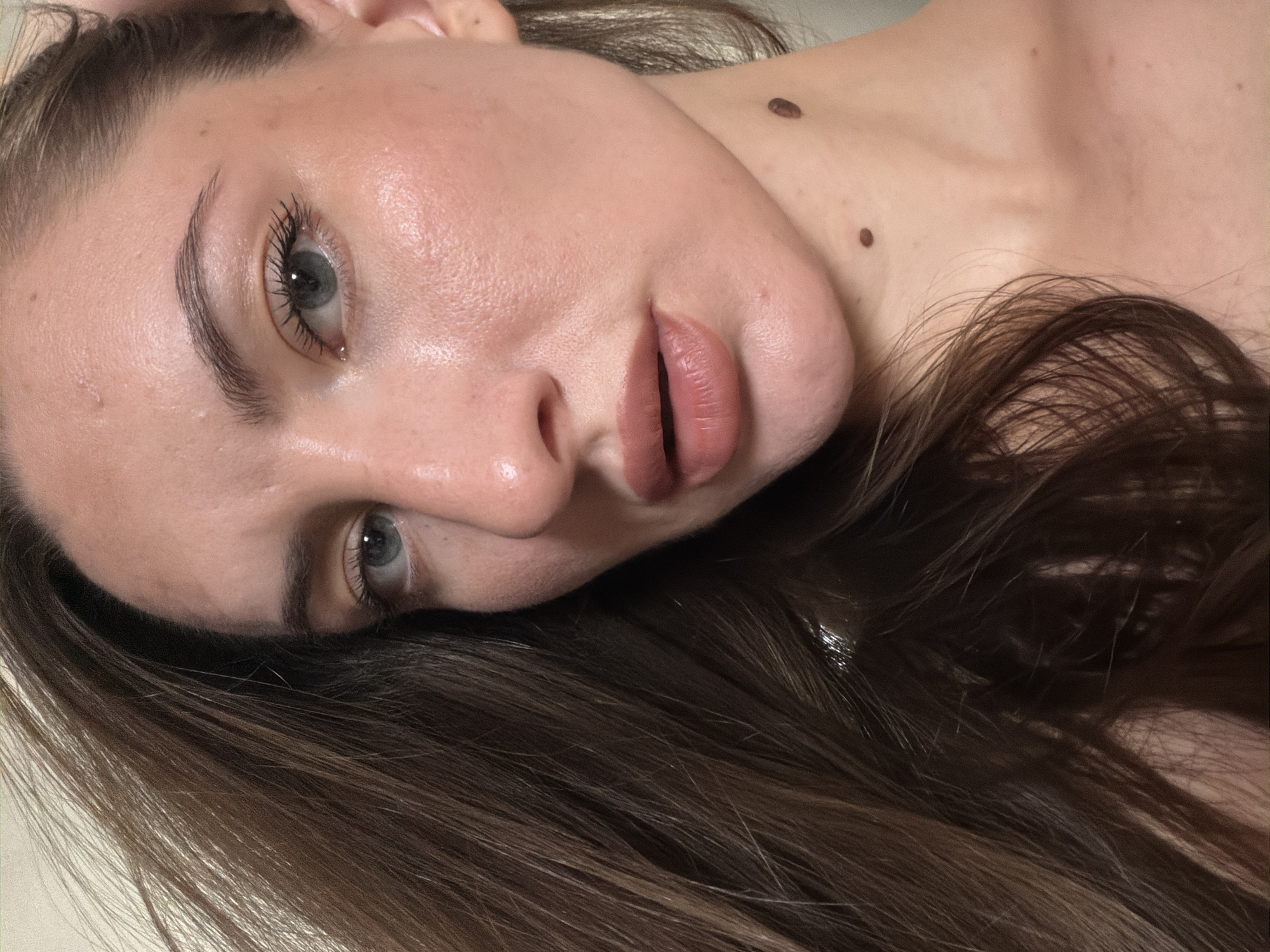 The image is a close-up, upward-angled shot of a person's face and upper chest, with their brown hair blurred and to the left of the frame. The person's skin appears fair and luminous, with some visible pores and a few small blemishes. Their light blue eyes are open and looking slightly to the left. Their eyebrows are well-defined and arched. The nose is prominent, and the lips are full and coated with a matte, nude-colored lipstick. There are two moles visible on the neck, one larger and darker than the other. The background is out of focus and appears to be a plain, neutral color. The overall lighting is soft and diffused, highlighting the contours of the face and creating a dewy effect on the skin. There is no indication of the city of Sharm El Sheikh, Egypt, in the image.