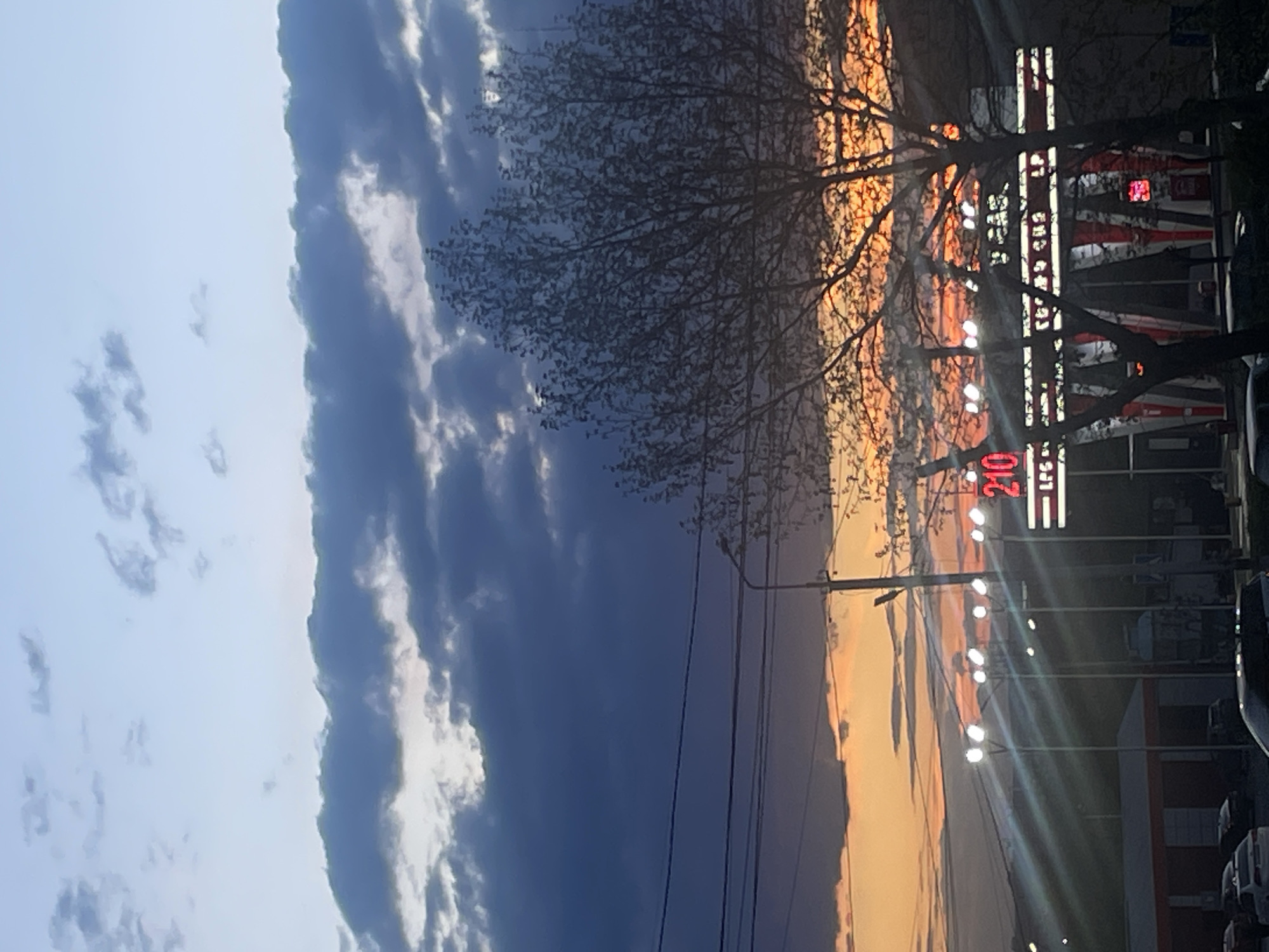 This image captures an urban or suburban scene at dusk, likely in Yerevan, Armenia, characterized by a dramatic sky and artificial illumination.
The sky dominates the upper portion of the frame, displaying a striking transition from light to dark. On the far left (top of the image if correctly oriented), the sky is a clear, light blue with scattered, wispy white clouds. Moving right, it transitions into a dense band of dark, heavy grey-blue clouds. Further to the right, closer to the horizon, the sky glows with vibrant orange and yellow hues, indicative of a sunset.
Silhouetted against this dynamic sky are the bare branches of a large deciduous tree, its intricate network of branches spreading across the middle of the frame. Power lines crisscross horizontally through the central sky, adding to the urban landscape.
The lower portion of the image (right side as oriented) reveals ground-level features. Several bright, circular streetlights or outdoor lamps are illuminated, casting strong rays of light. A prominent, vertical commercial sign stands out, featuring red illuminated numbers "240" and white letters "LFC". Faint outlines of buildings or structures are visible behind these lights and the tree, along with partially visible parked vehicles on the far right and bottom.
The combination of the setting sun, heavy clouds, and active artificial lighting strongly suggests the time of day is dusk or early evening. The scene conveys a sense of transitioning light and the start of night in a developed area.