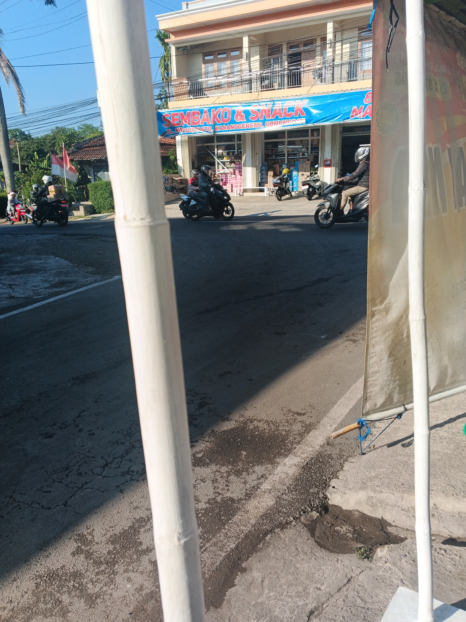 The image depicts a street scene in Semarang, Indonesia, with a two-story building housing a "SEMBAKO & SNACK" store. Several people are visible on motorcycles, with some riding and others parked. The store's facade features a large blue banner with white lettering. In the foreground, a white bamboo pole partially obstructs the view, and a rolled-up banner is visible on the right side of the frame. The street is paved and shows signs of wear and tear. A red and white Indonesian flag is also visible, attached to a pole on the left side of the image. The overall scene suggests a commercial area with moderate traffic and pedestrian activity.