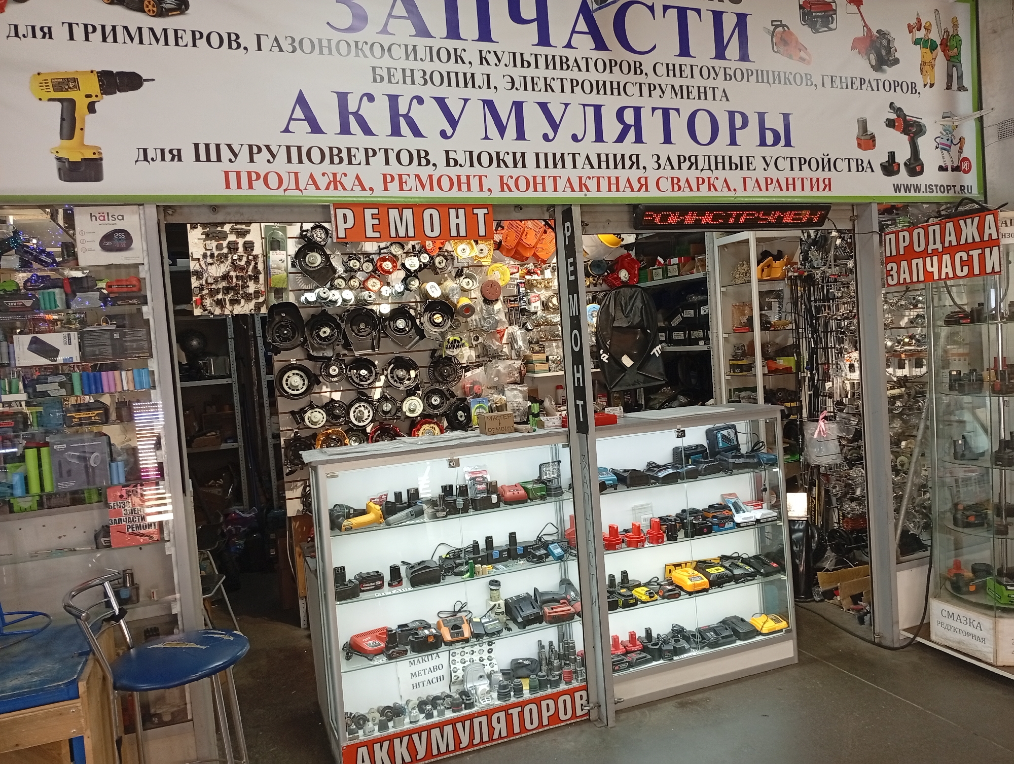 The image shows the storefront of a shop selling power tool parts and accessories. A large sign above the entrance displays "ЗАПЧАСТИ" (Spare parts) and lists various tools and equipment such as trimmers, lawn mowers, snow blowers, generators, chainsaws, and electric tools. Below this, it reads "АККУМУЛЯТОРЫ" (Batteries) and mentions sales, repair, and warranty for battery chargers, power supplies, and welding equipment. A smaller orange sign in the center reads "РЕМОНТ" (Repair).
The shop interior is visible through glass display cases filled with batteries, chargers, and other power tool components. To the right, another section of the shop has more shelves stacked with various parts. In front of the shop, a blue and chrome bar stool is placed near a wooden stand. The overall impression is that of a retail environment specializing in tools and their associated parts, located in an urban setting.