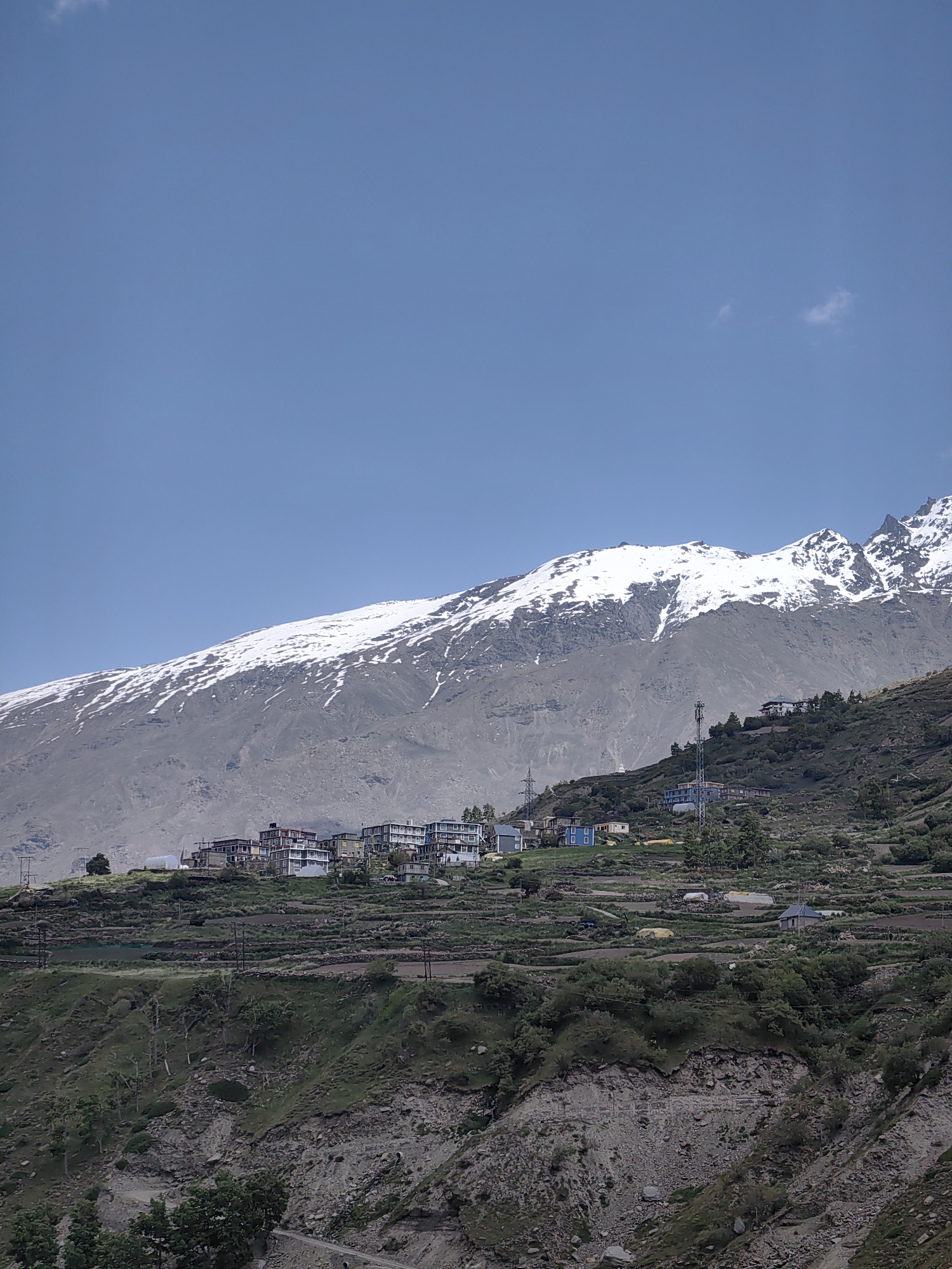 The image shows a village nestled on a mountainside in India. The village features a cluster of buildings with various architectural styles, some appearing to be multi-story structures with balconies. In the background, a snow-capped mountain range dominates the skyline, with patches of snow clinging to the rugged slopes. The terrain in front of the village is terraced, suggesting agricultural activity, with greenery and some scattered trees. A cell tower is visible on the hillside, indicating modern infrastructure within the rural setting. The sky is clear blue with a few wispy clouds.