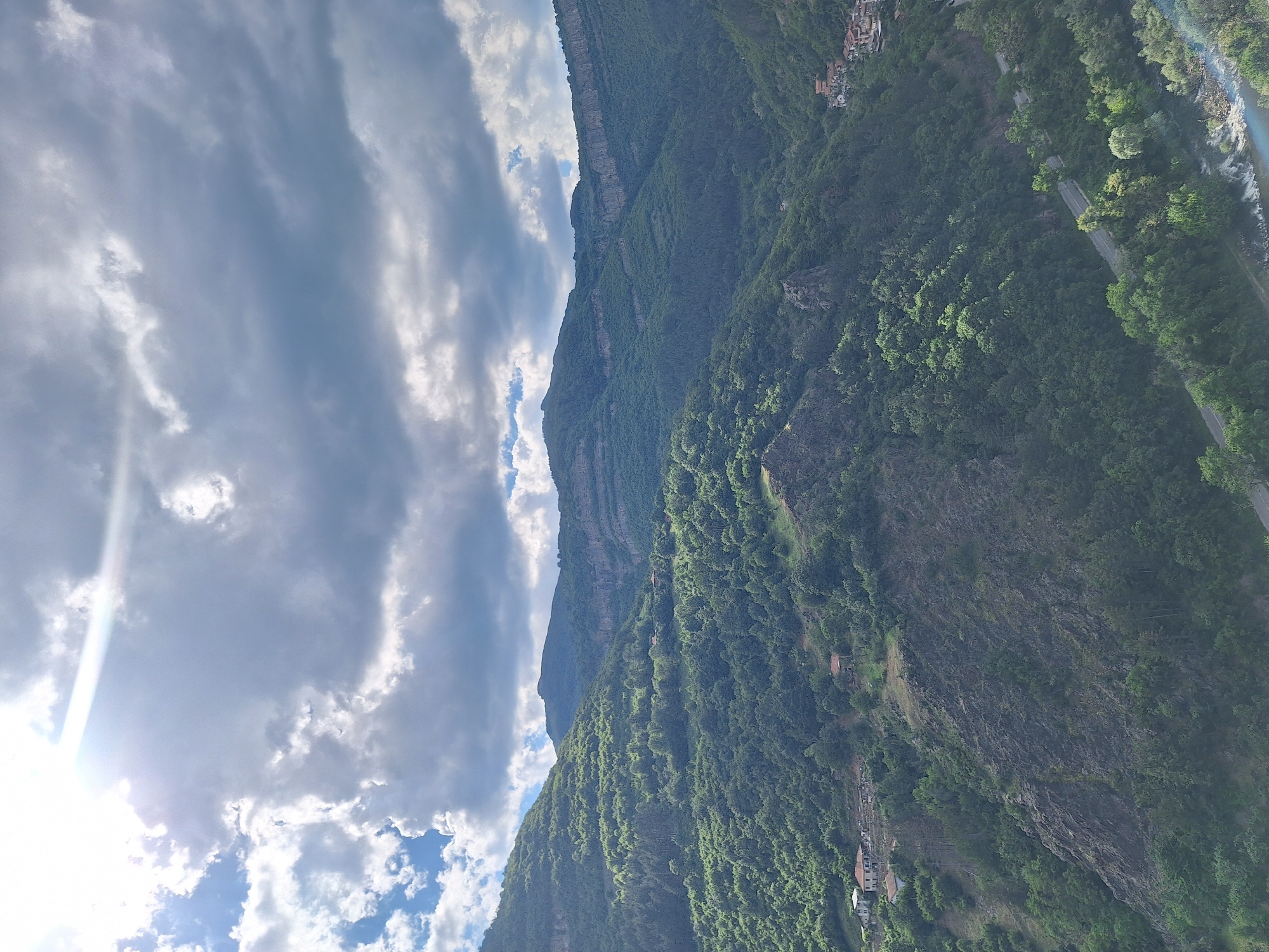 The image captures a view of mountainous terrain, with a section of the city of Zasele, Bulgaria visible on the right side of the image. A river and road are visible running alongside the base of the mountain. The mountain itself is densely covered in green foliage, with visible rock formations and some small structures dotted across its surface. The sky is filled with a mix of white and grey clouds. The sun is visible in the lower left corner of the image, creating a bright glare.