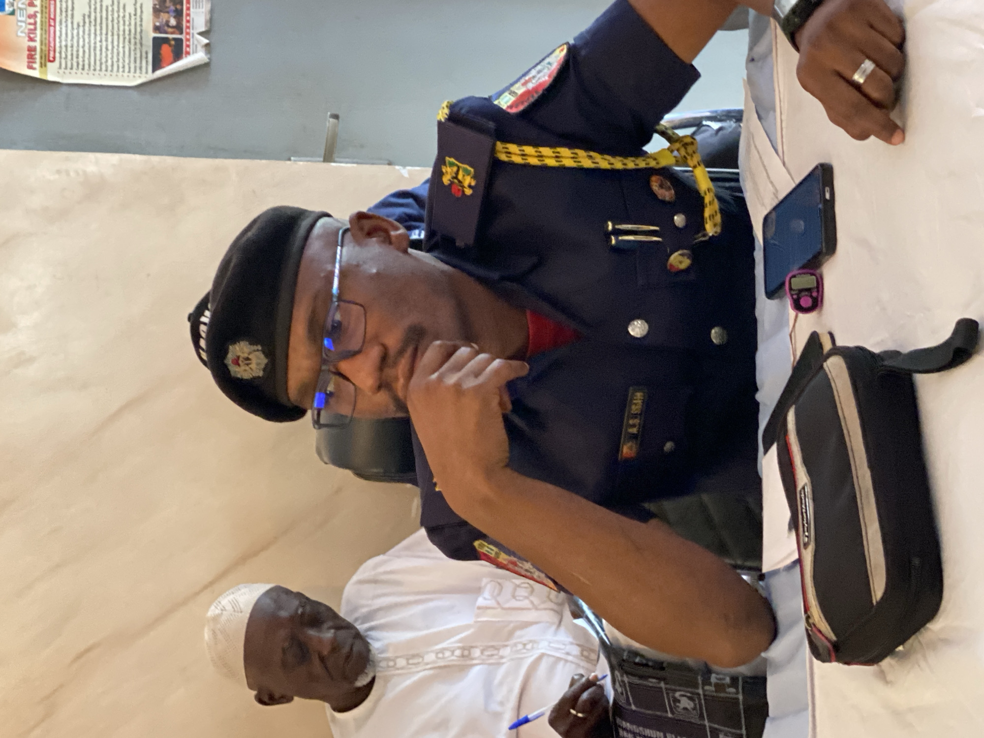 In the city of Unguwar Sarki, Nigeria, a middle-aged Black man wearing a white kufi cap and traditional white attire is seated, looking downwards with his head tilted. To his right, a younger Black man in a dark blue uniform with a beret is seated, his hand resting on his chin as he looks to the side. The uniform has insignias on the collar and epaulets, and a yellow cord hangs from his right shoulder. A white name tag on his uniform reads "A.S. USMAN". On the table in front of them are various items, including a dark bag with red and white trim, a cell phone, a pink digital counter, and a pen. Behind the seated men, a portion of a poster with text about fire safety is visible on the wall. The environment appears to be an indoor setting, possibly an office or meeting room, with light-colored walls.