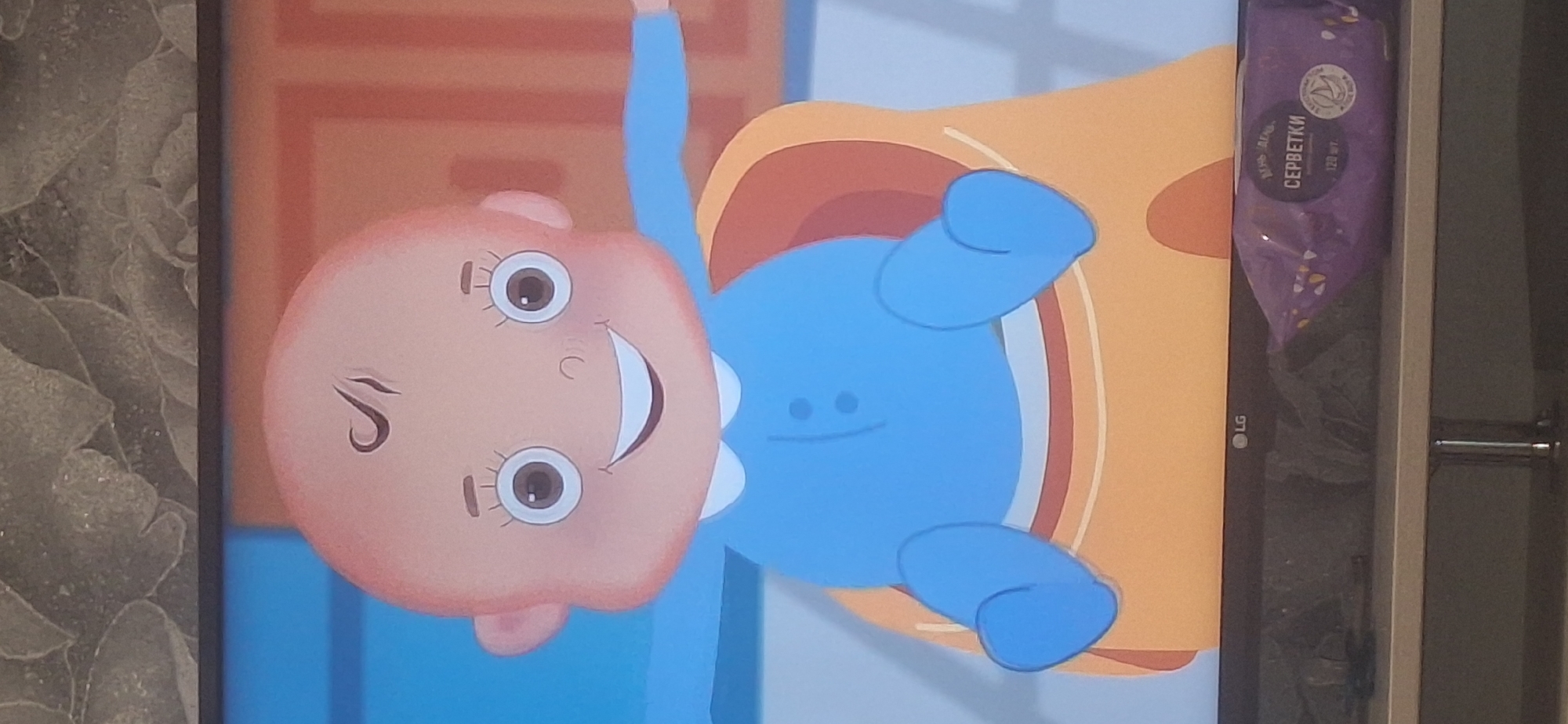 A cartoon baby is depicted sitting in a large orange bowl against a blue and orange background. The baby has a bald head with a single swirl of hair, large round eyes with eyelashes, and a wide smile. It is wearing a blue onesie with a white collar and two dark buttons. The baby is positioned in the center of the frame, with its legs and arms visible. In the foreground, on a shelf below the screen, is a purple package of wet wipes labeled in Russian. The LG logo is visible on the bottom edge of the screen. The image appears to be a screenshot taken from a television screen, with a textured grey background visible at the top. There is no indication of the location in Bila Tserkva, Ukraine.
