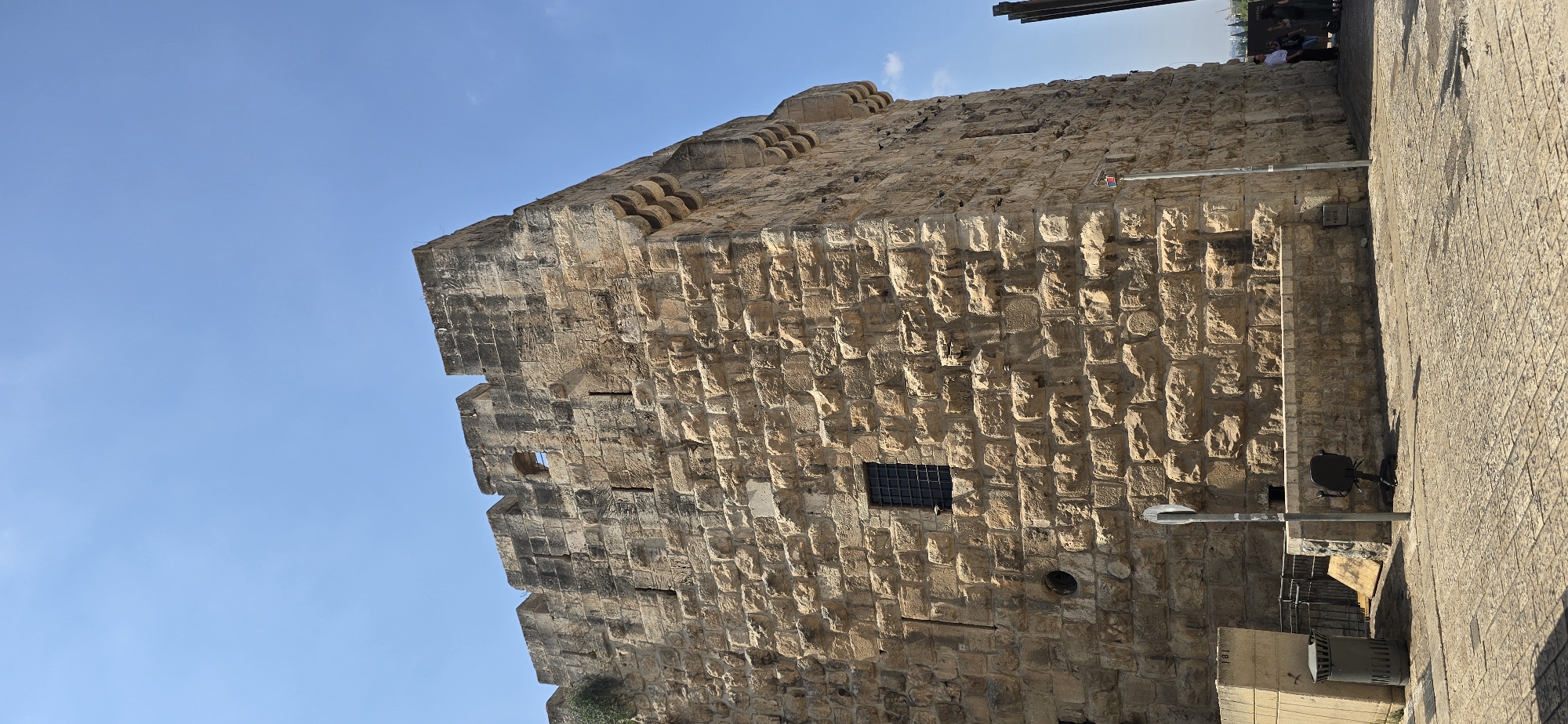 The image captures a section of the ancient stone walls of Jerusalem, Israel, under a clear blue sky. The wall is constructed from large, roughly hewn stone blocks, exhibiting a weathered texture and varying shades of tan and brown. Along the top of the wall are battlements, and a small, barred window is visible in the stone facade.
In the foreground, a paved cobblestone area leads up to the wall. To the left, a grey metal trash receptacle stands next to a low stone barrier. A black office chair is positioned on the cobblestones in front of the wall. A metal pole with a circular sign is present. Further to the right, at the edge of the frame, a dark vertical structure, possibly a fence post or part of a sign, is visible. A few people are seen in the distance on the right, walking near the wall. The overall impression is one of historical significance and weathered grandeur.